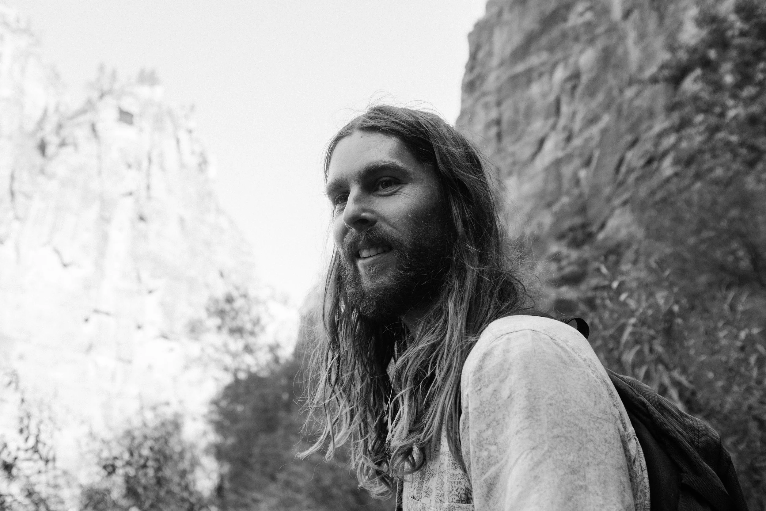 A man with long hair and beard smiling outdoors with a rocky cliff and trees in the background.