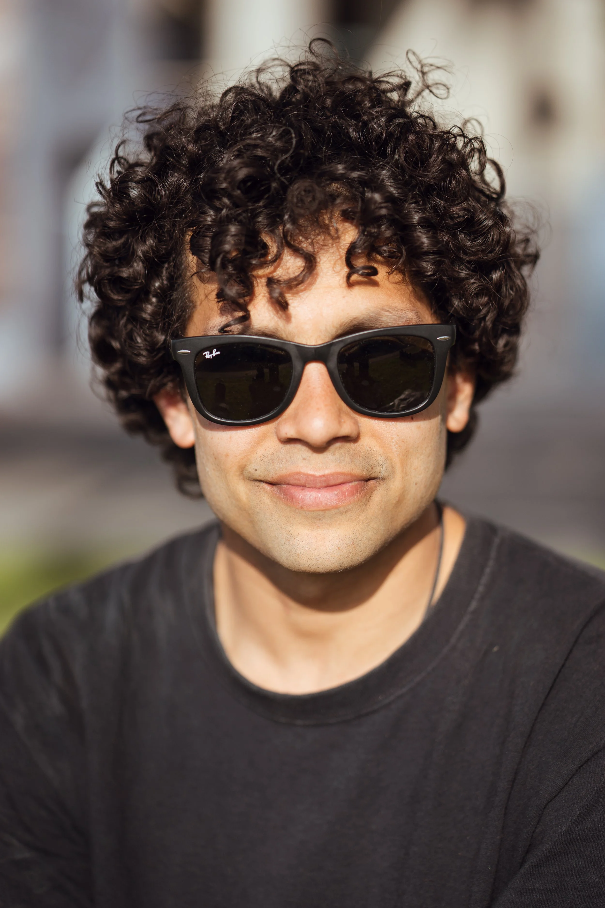 A man with curly dark hair wearing black sunglasses and a black t-shirt, smiling outdoors in bright sunlight.