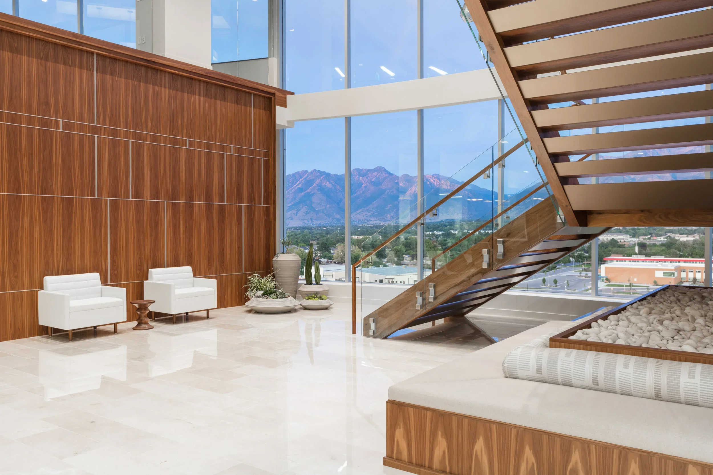 Modern lobby with large glass windows showing mountain view, white seating area, wooden wall, staircase with glass and wood railing, decorative plants, and a clear bright sky.