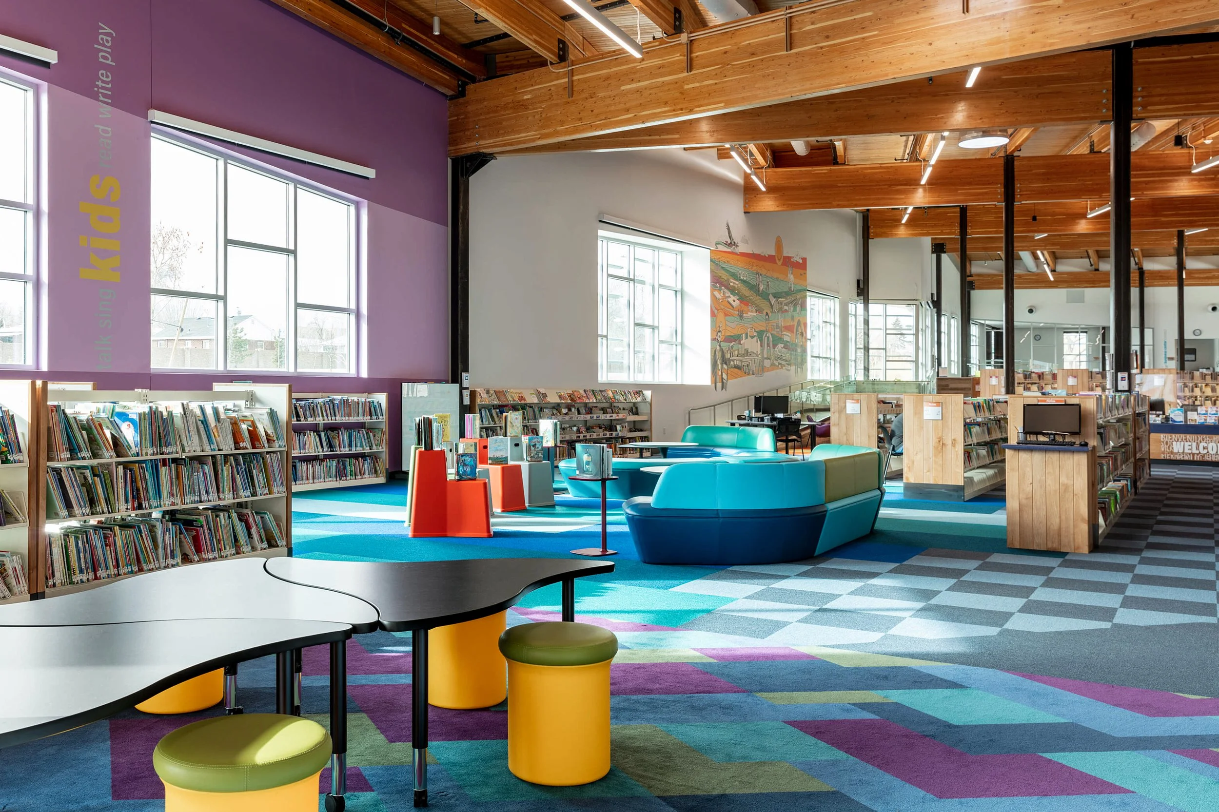 A colorful library interior with bookshelves, a reading and seating area with modern furniture, large windows, and light wood ceiling beams.