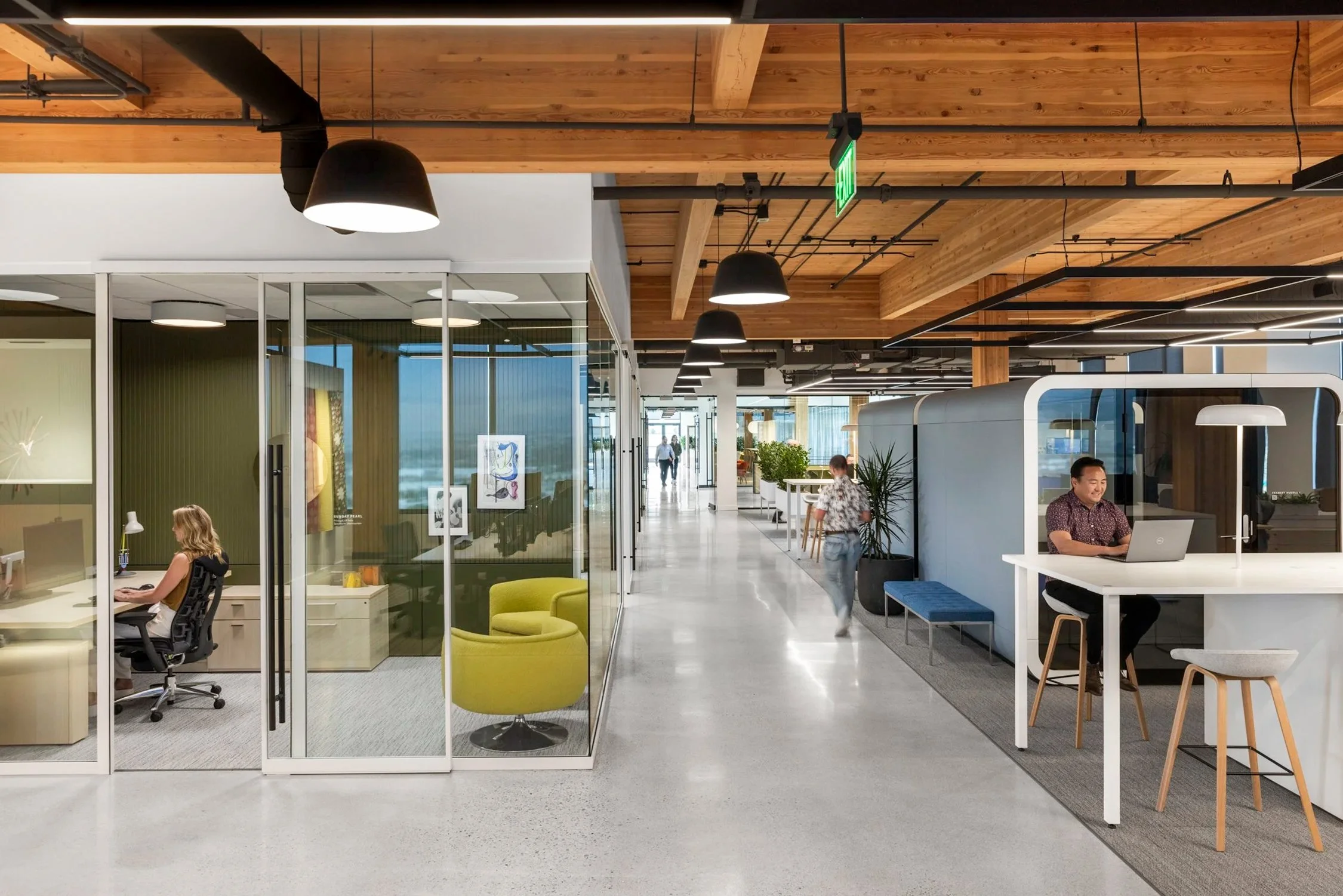 Modern office lobby with glass private rooms, open seating area, people working at desks and walking, wooden ceiling, black pendant lights, exit sign.