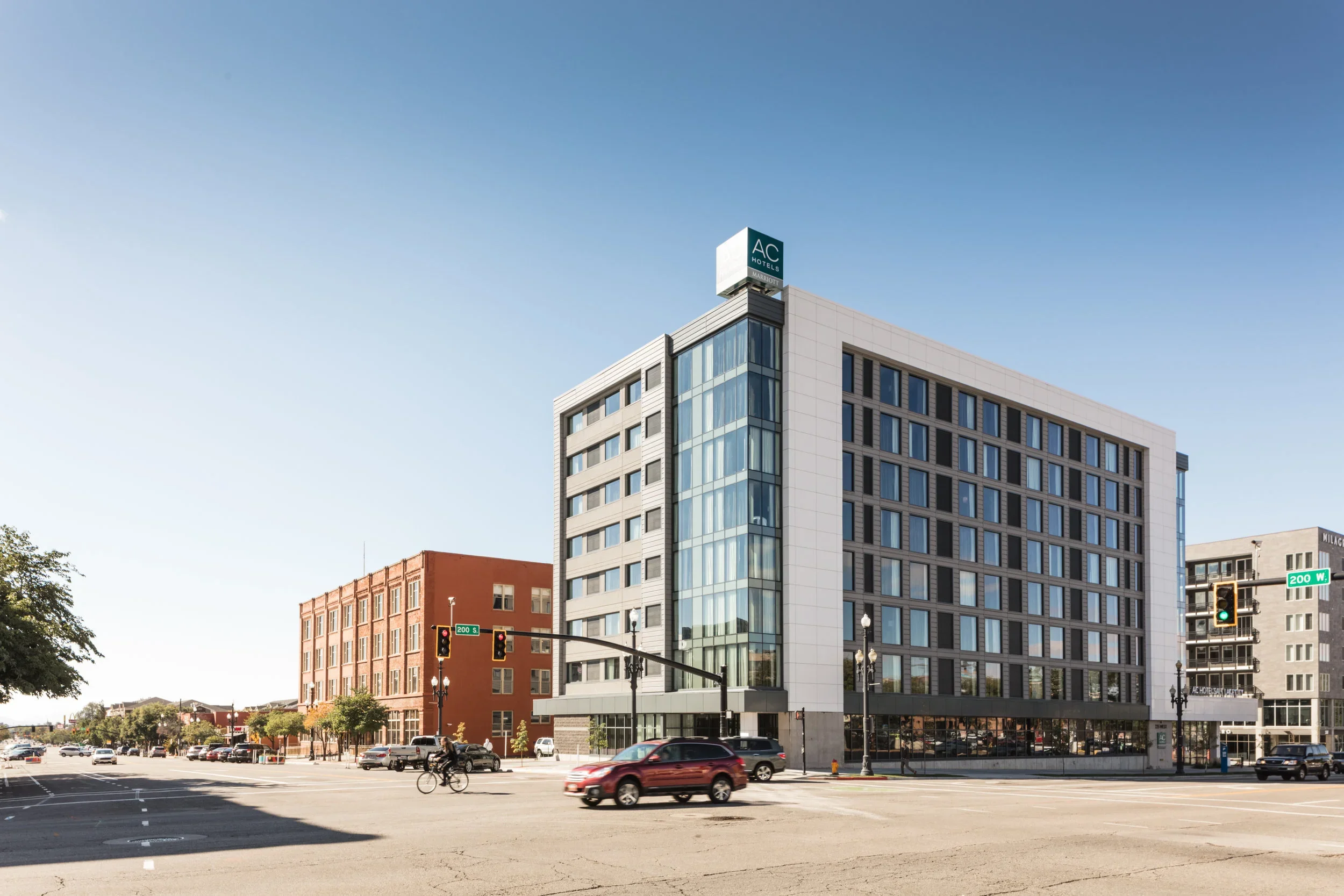 A city street with a modern multi-story hotel building labeled 'AC Hotels by Marriott' at the corner of 200 W and 200 S streets. Cars are parked and moving along the street, and a cyclist is riding across the intersection. The sky is clear and blue.
