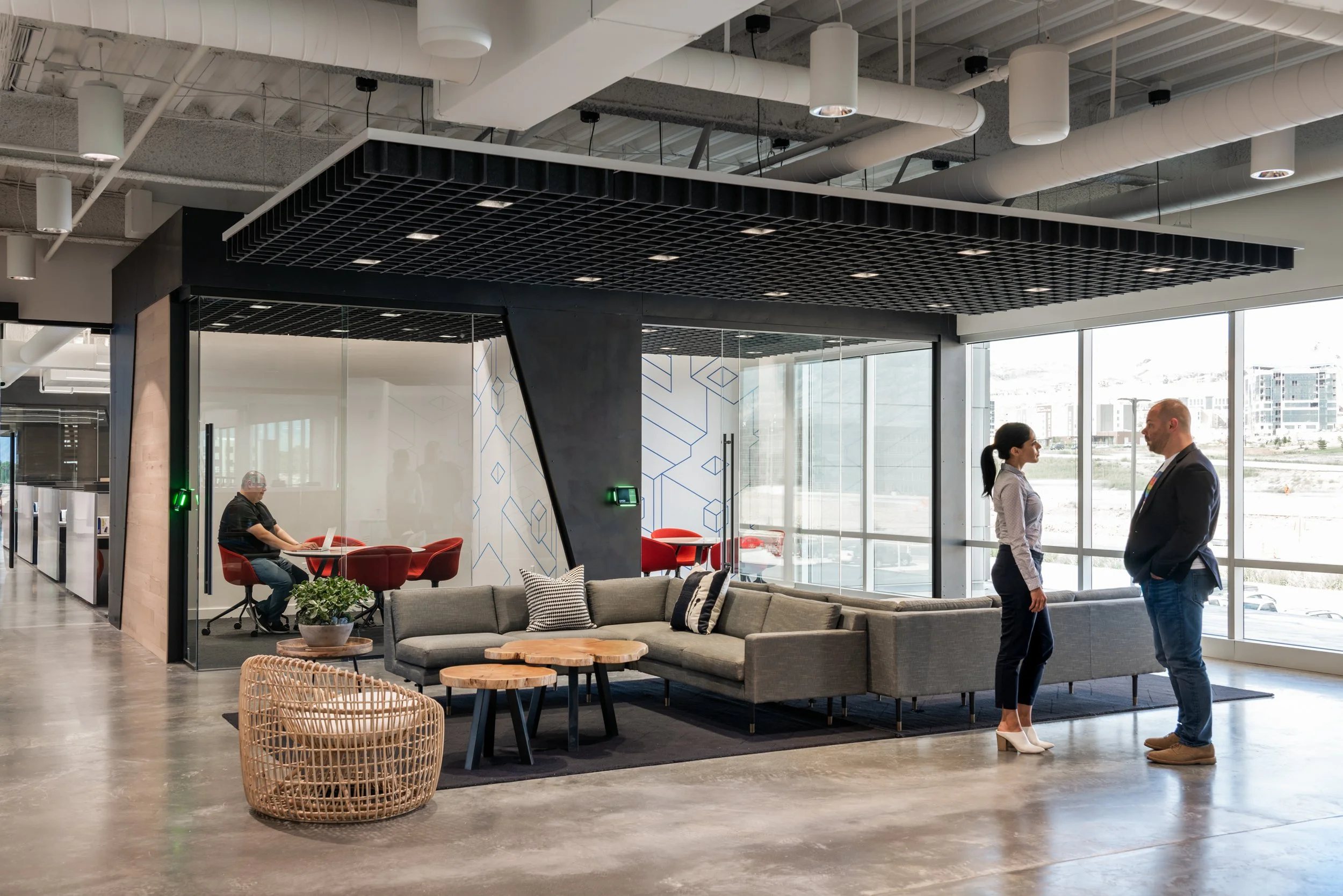 Modern office reception area with large windows, gray sofas, red chairs, glass wall, and a ceiling with black grid panels and white ducts. Two people are standing and talking, a woman in white heels and a man in casual attire with a colored lanyard. 