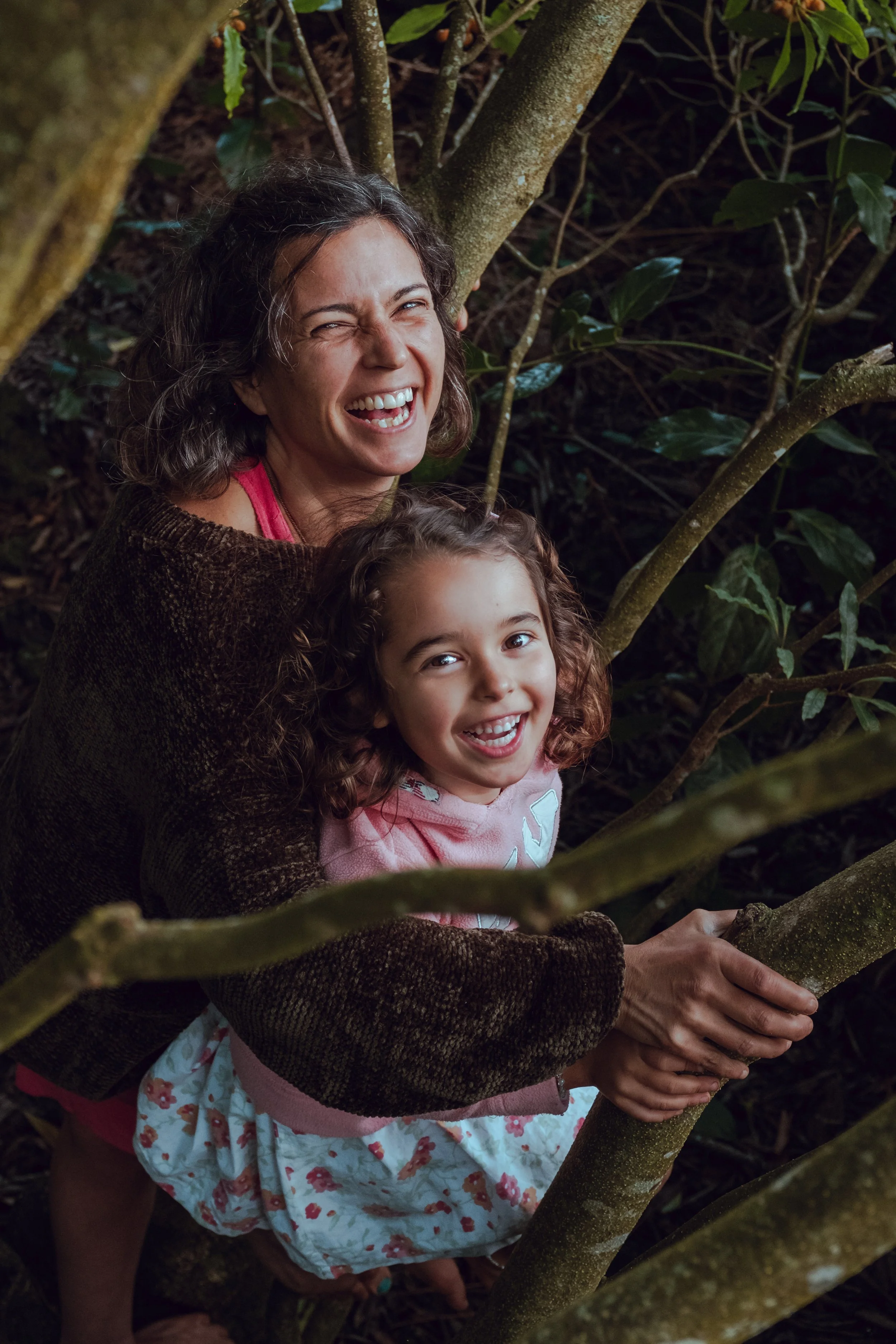A woman and a young girl smiling and hugging among tree branches with green leaves.