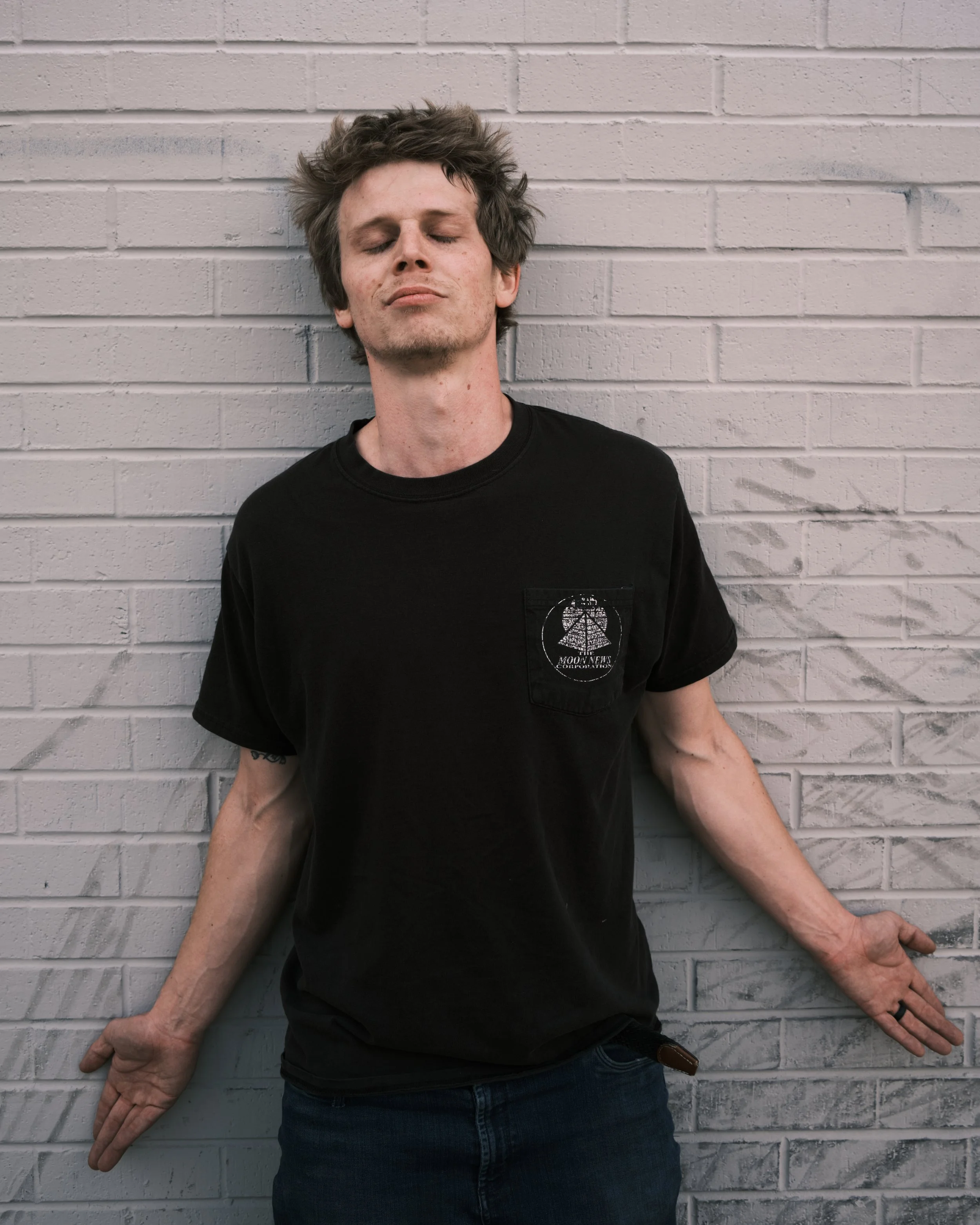 A young man with messy hair leans against a white brick wall with his eyes closed and arms outstretched.