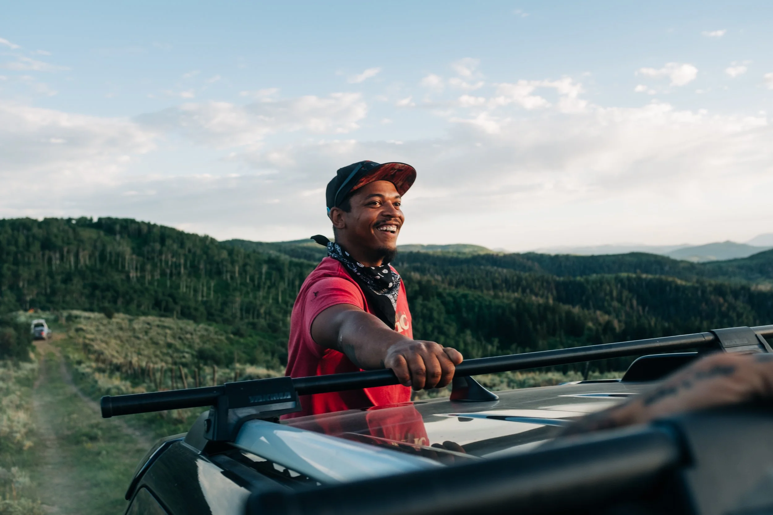 A young man smiling and riding on the roof of a vehicle in a rural, hilly landscape with green trees and a partly cloudy sky.