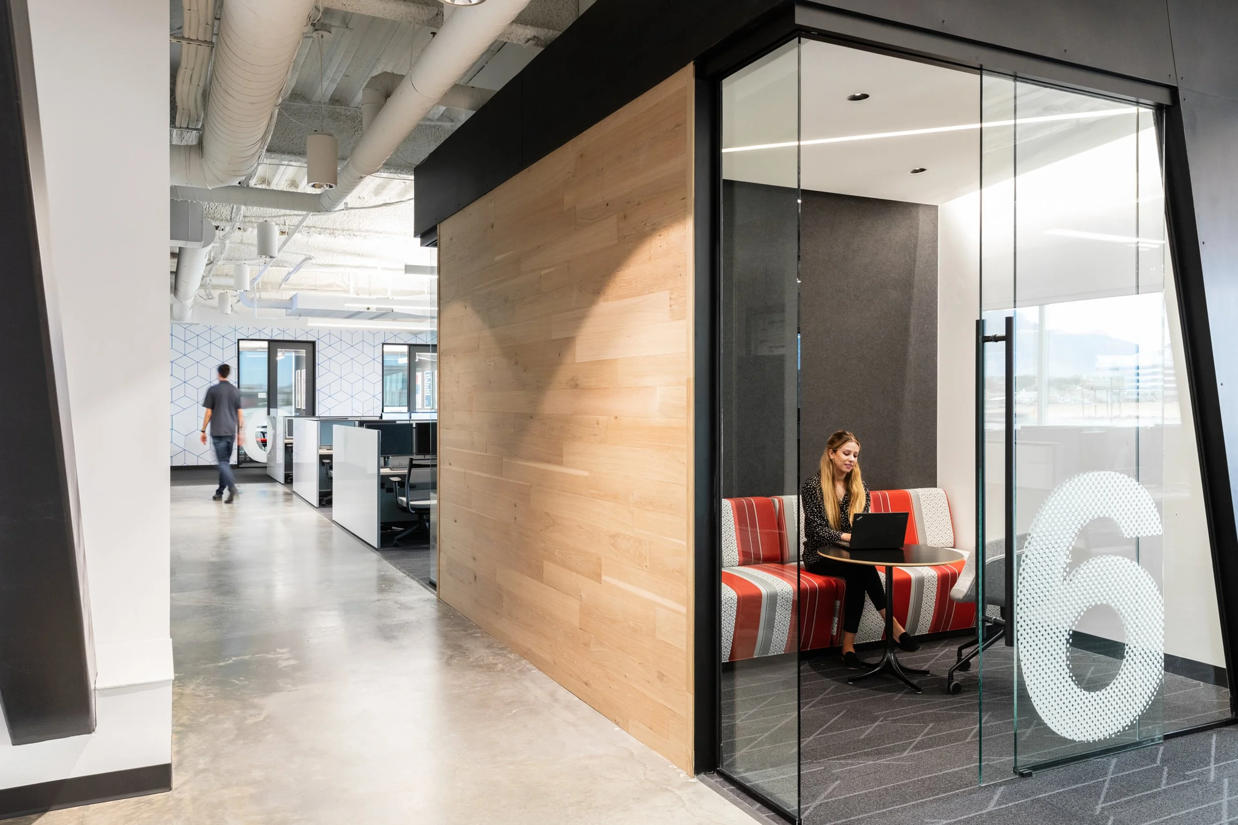 Modern office space with glass-walled meeting room, wooden partition, and open workspace with desks and chairs. A woman is working on a laptop inside the glass room, and a man is walking through the office in the background.