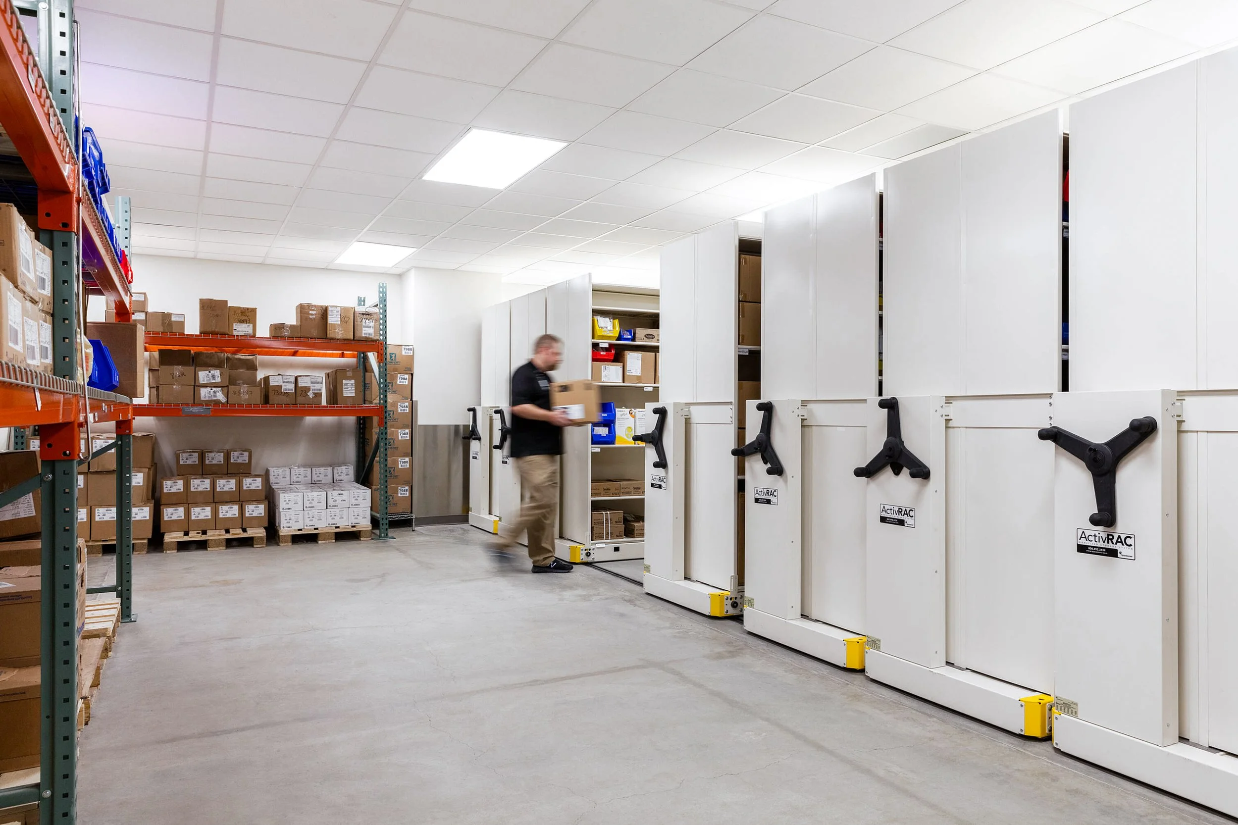 Warehouse with shelves full of boxes and a worker operating a pallet wrapping machine.