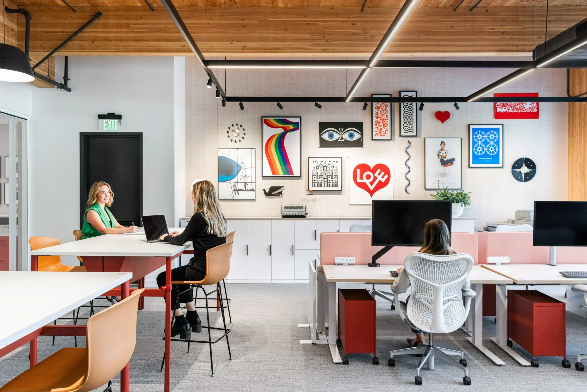 An interior office space with white walls, wooden ceiling, and modern decor. There are several women sitting at desks, working on laptops. The wall features framed artwork, including a rainbow, a large red heart with 