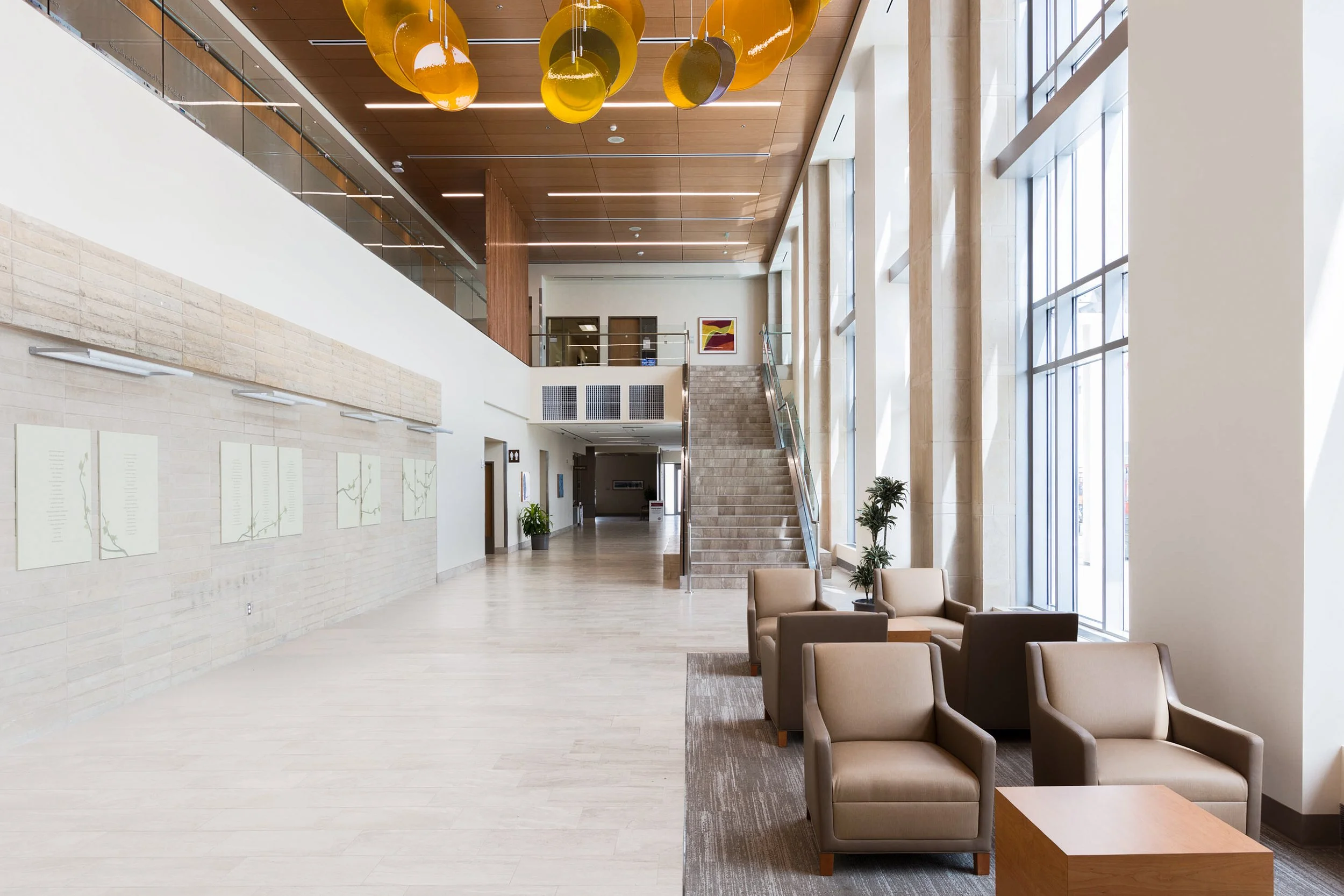 Modern lobby with beige armchairs, a wooden coffee table, large windows with natural light, a staircase, abstract wall art, and yellow hanging light fixtures.