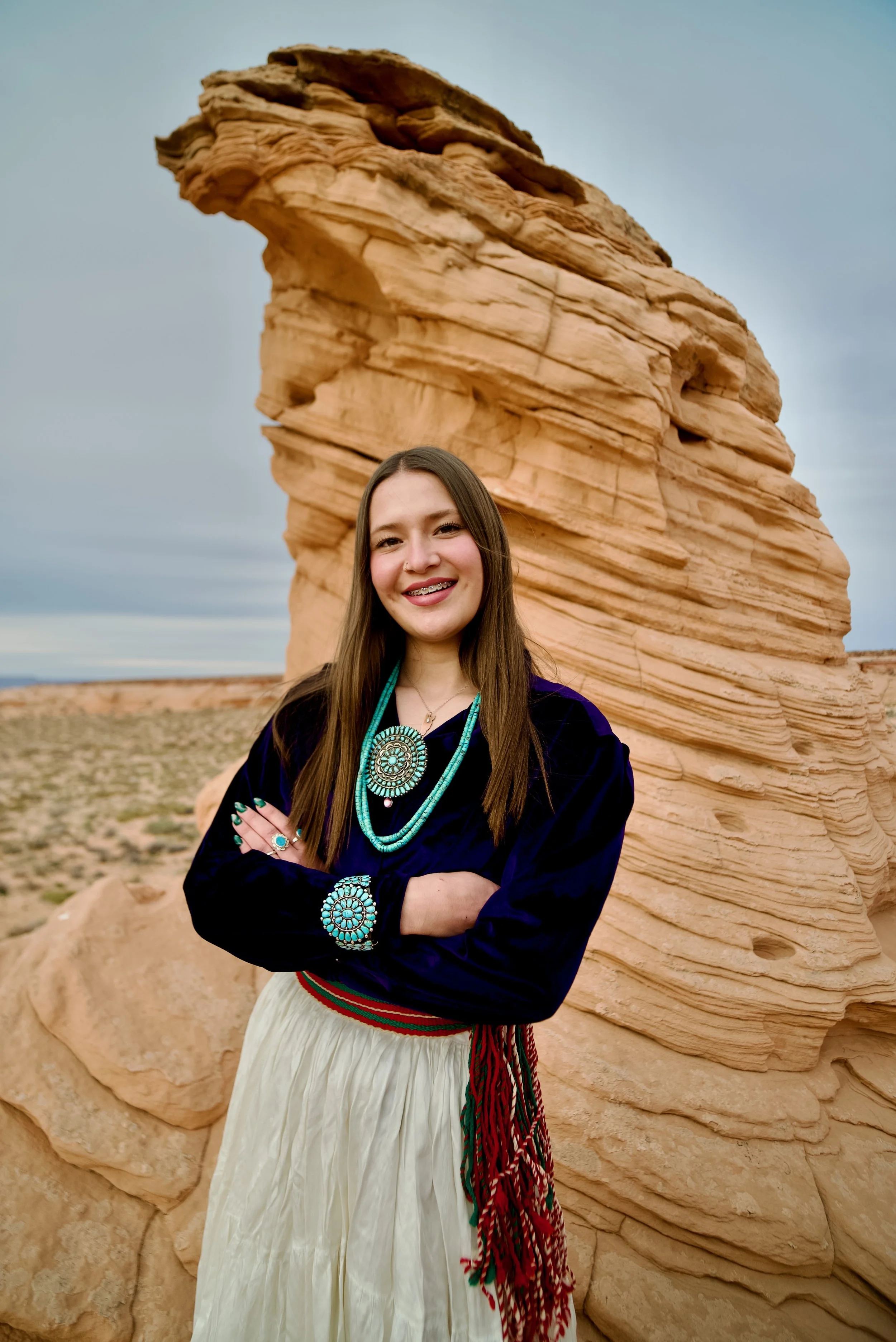 Young woman standing in front of a large rock formation, smiling and wearing traditional Southwestern jewelry and clothing.