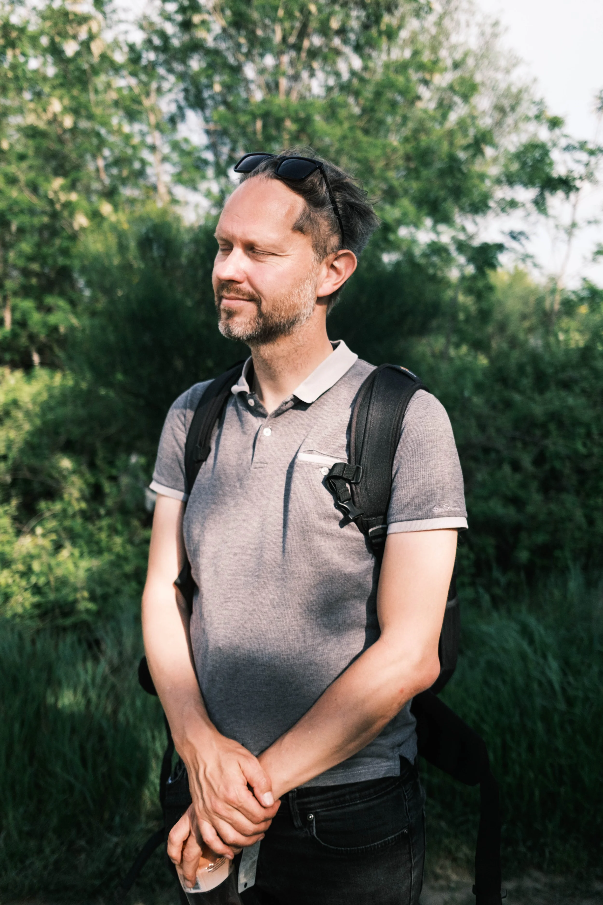 A man with a beard, gray polo shirt, and backpack standing outdoors with closed eyes and a peaceful expression.