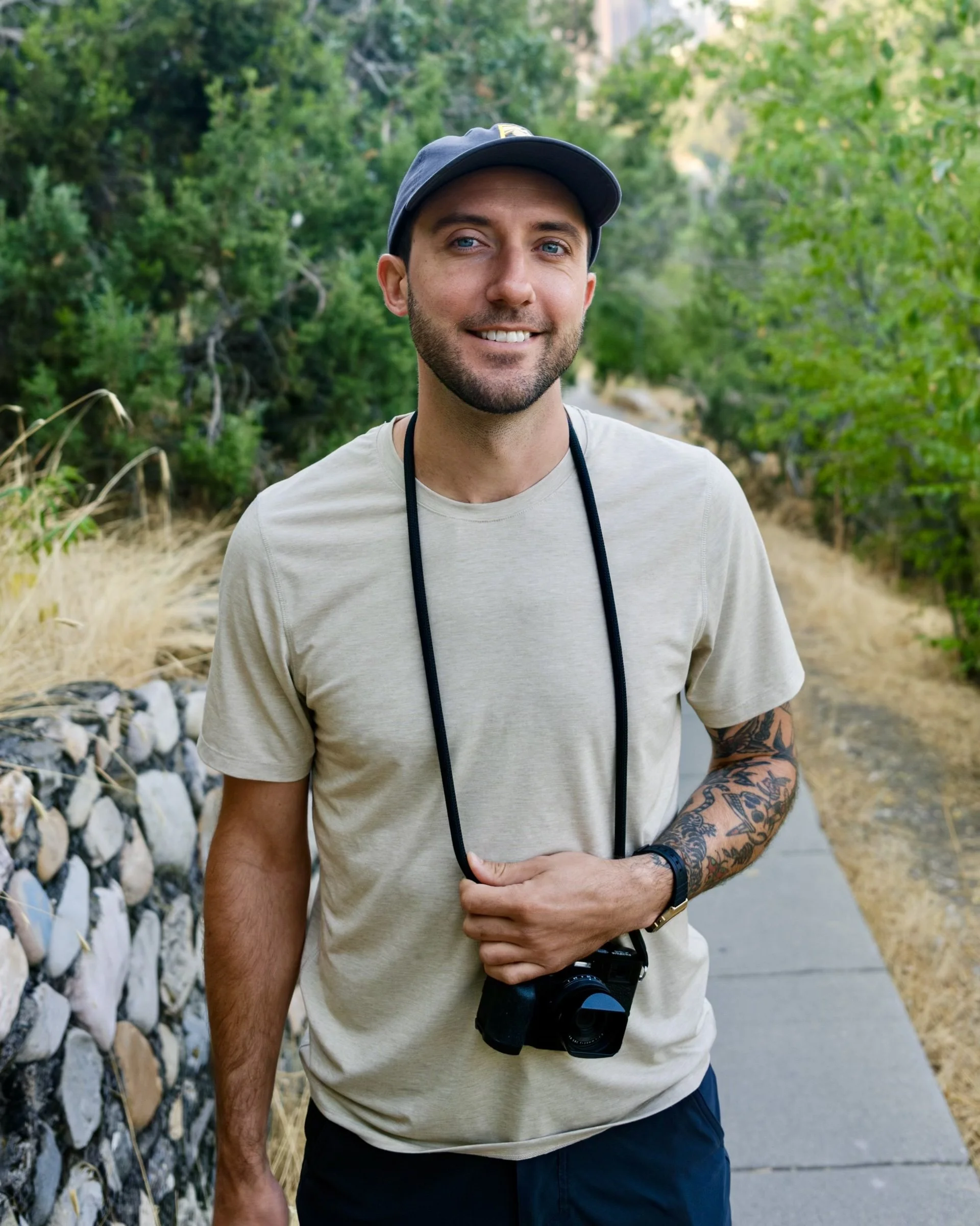 A young man with tattoos on his left arm, wearing a beige t-shirt, a black watch, and a black baseball cap. He is holding a camera in his left hand and a camera strap around his neck, standing outdoors in a green, wooded area.