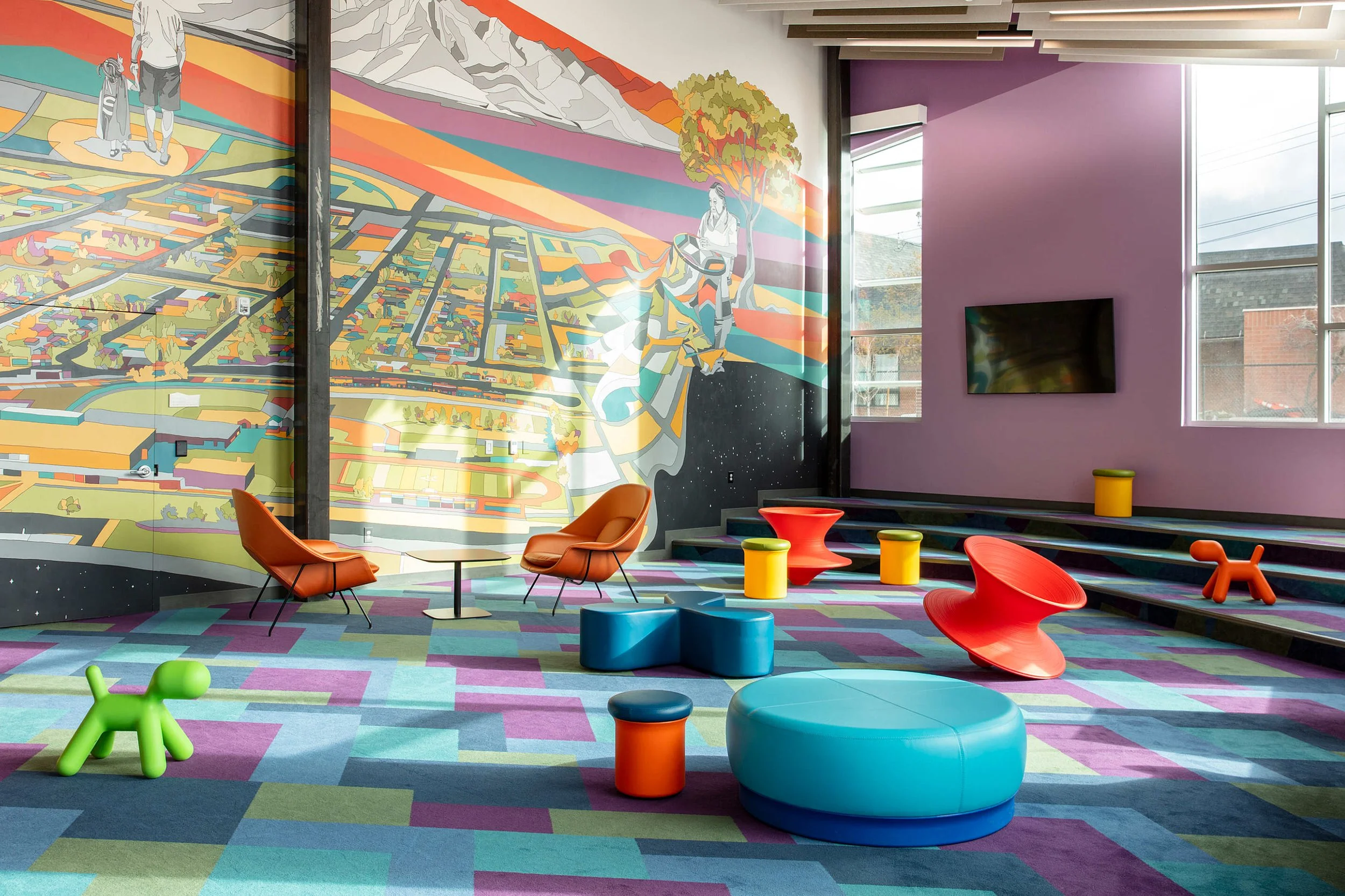 Bright and colorful interior space with abstract geometric patterned carpet and modern furniture, including orange chairs, a blue circular bench, and small stools in various colors. A large mural on the wall depicts a stylized landscape with mountain