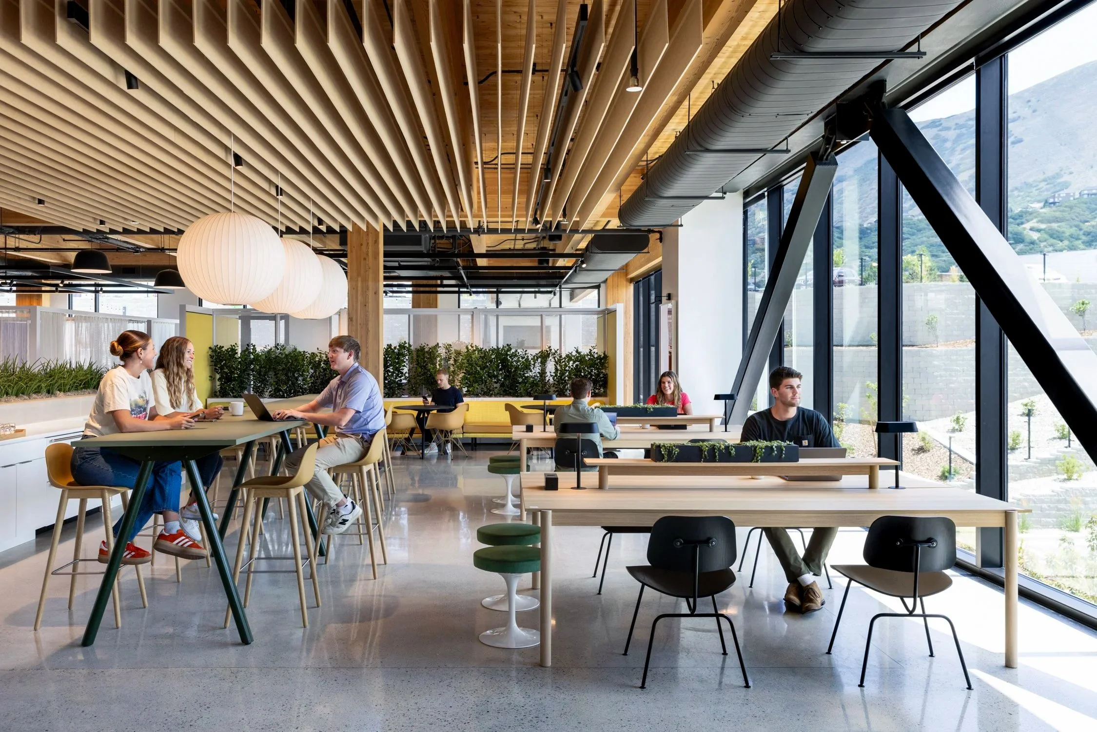 Modern office or co-working space with large windows, wooden ceiling and beams, and contemporary furniture. Several people are working and chatting at desks, with some using laptops and others on their phones. The room has natural light and a mountai