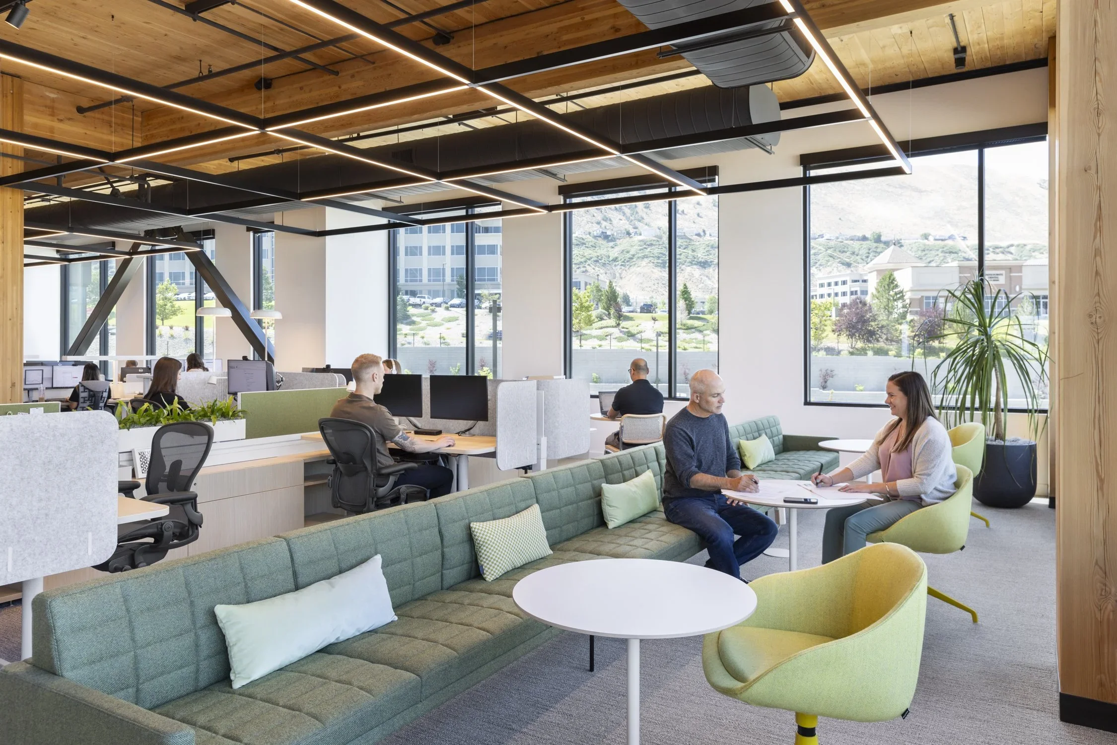 Modern office space with large windows, wooden ceiling, and abstract black and orange lighting fixtures. People are seated at desks working on computers, and some are engaged in conversation at small tables with green and yellow chairs.