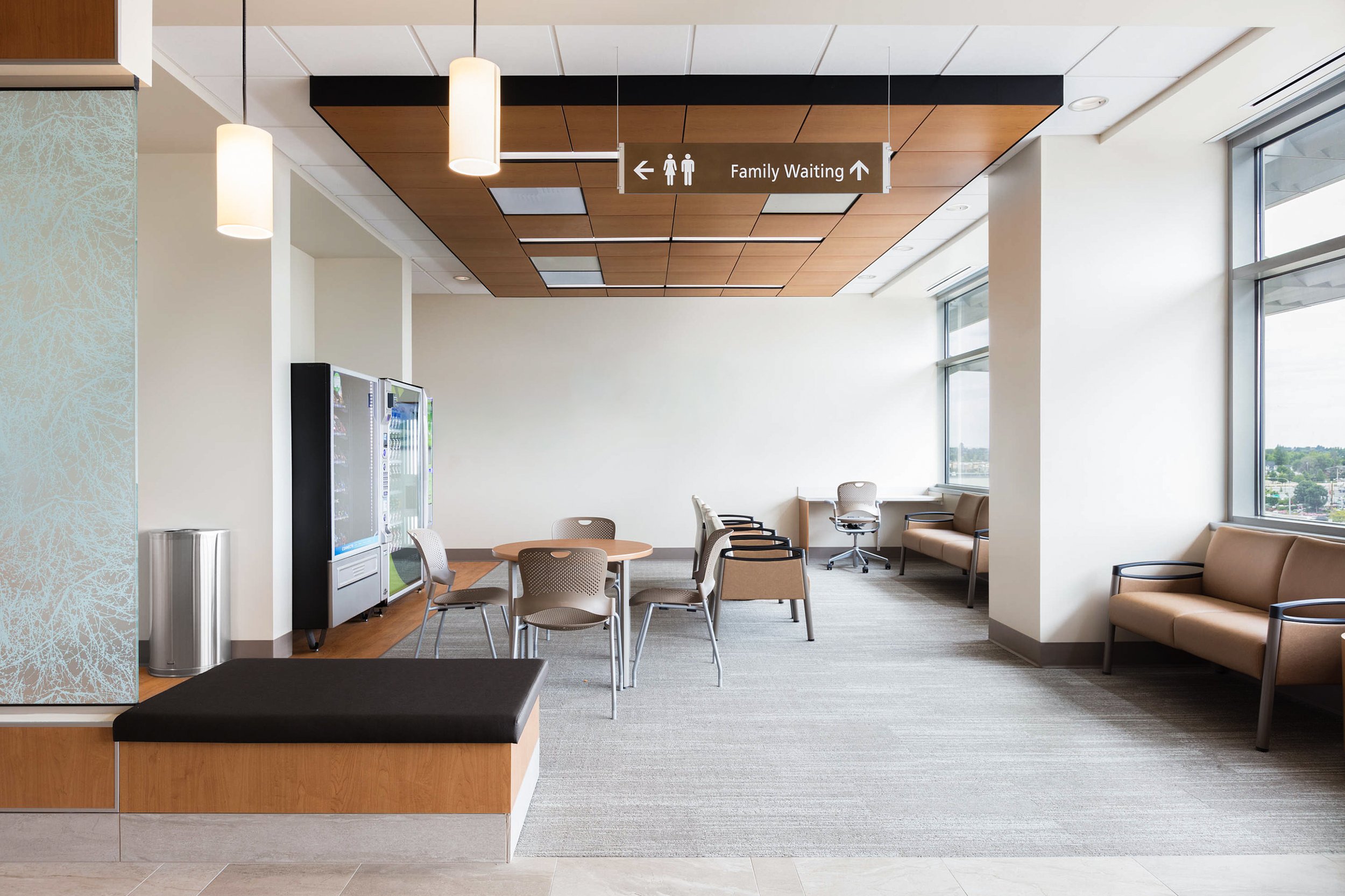 Interior view of a healthcare waiting area with chairs, tables, a vending machine, and a sign for family waiting with directions to restrooms.