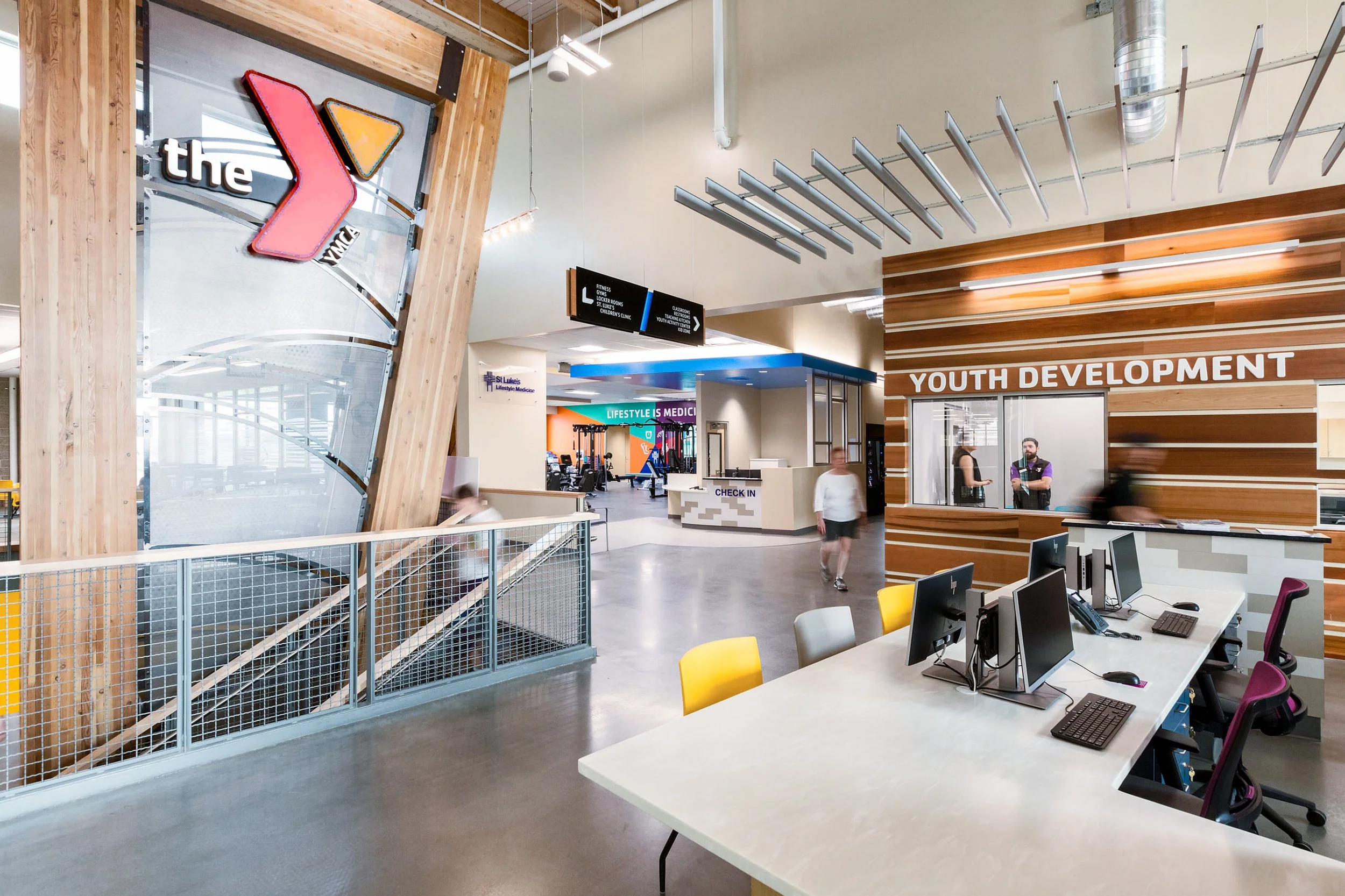 Interior of a YMCA facility with a reception desk, a sign pointing to fitness, locker rooms, and children's clinic, and a designated area for youth development.