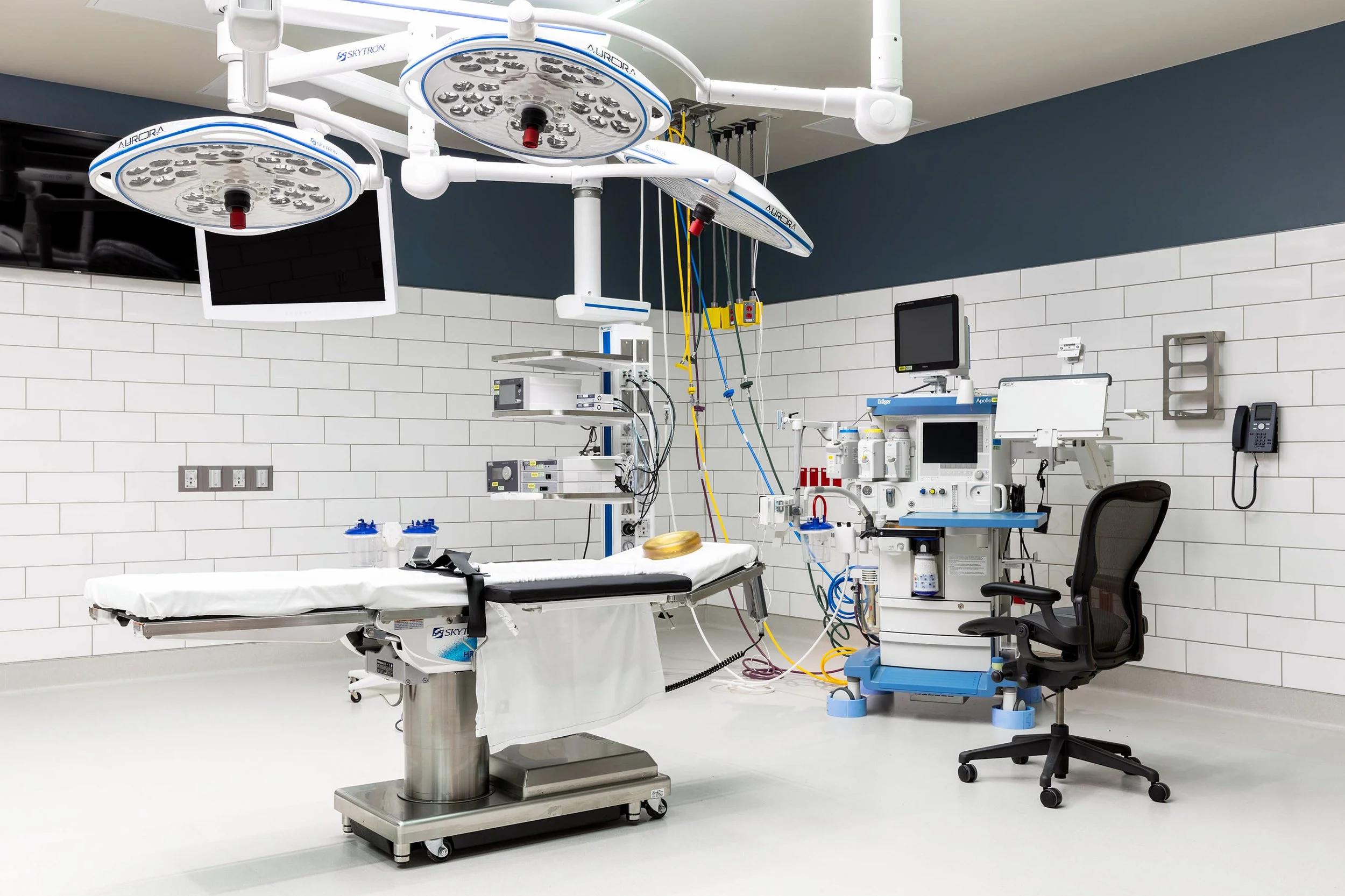 Medical operating room with surgical lights, hospital bed, medical monitors, and various medical devices and equipment.