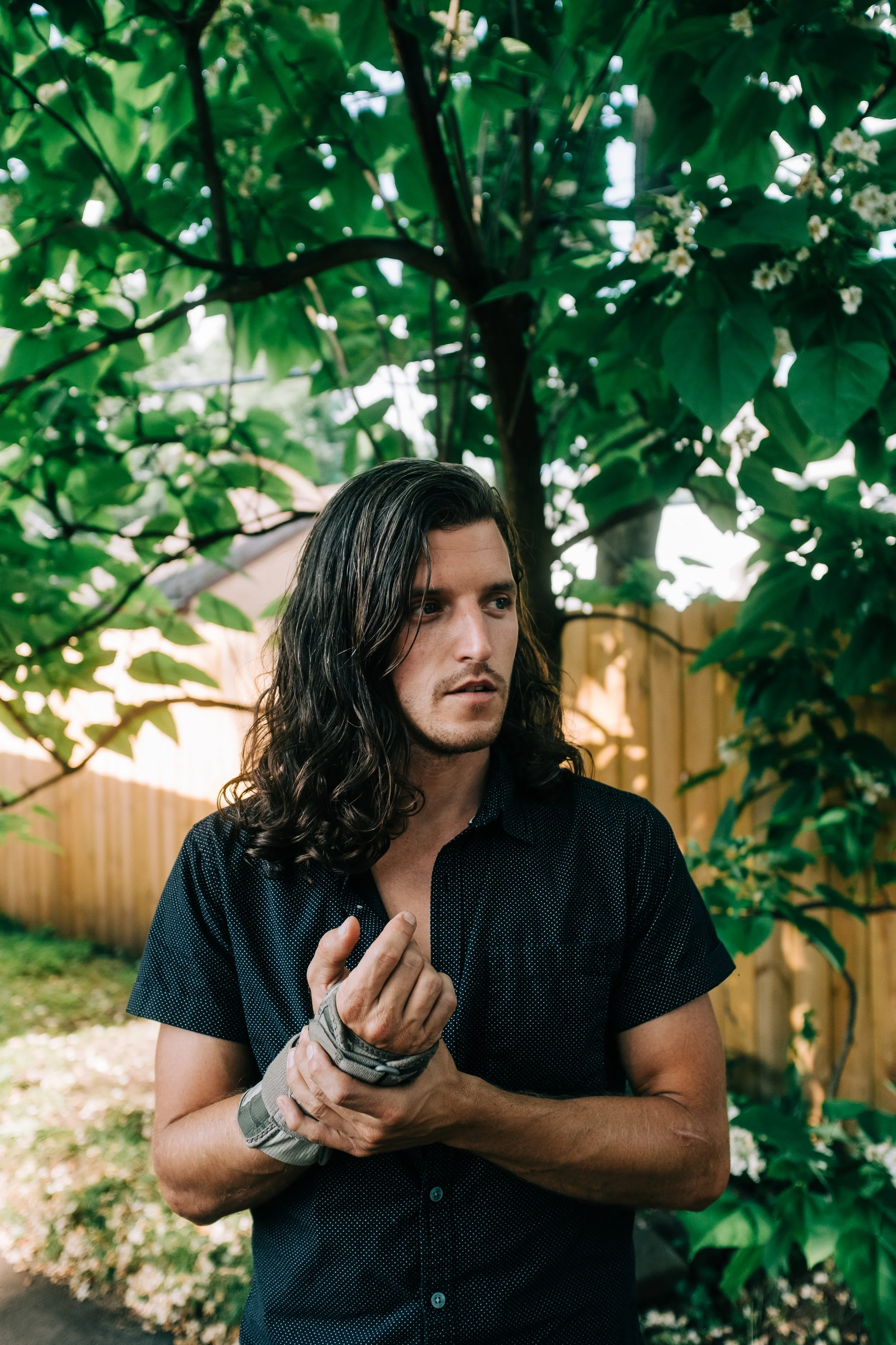 A young man with long, dark, wavy hair stands outdoors near a tree with green leaves, holding his hands together and looking to his right with a serious expression. A wooden fence is in the background.