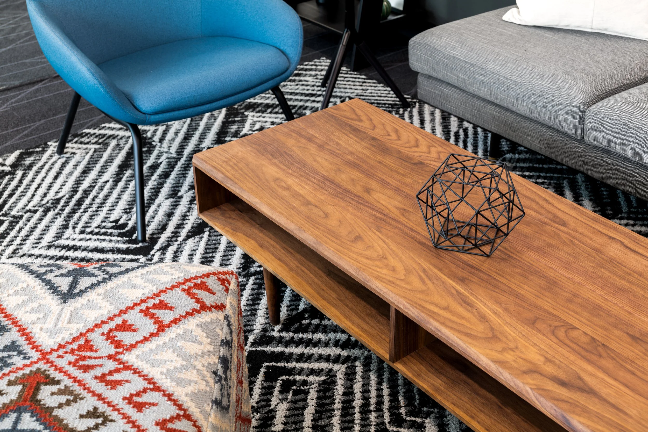 A living room with a blue chair, a gray sectional sofa, a wooden coffee table with a black wire geometric sculpture, a patterned black and white rug, and part of a patterned pillow and an armrest of the sofa.