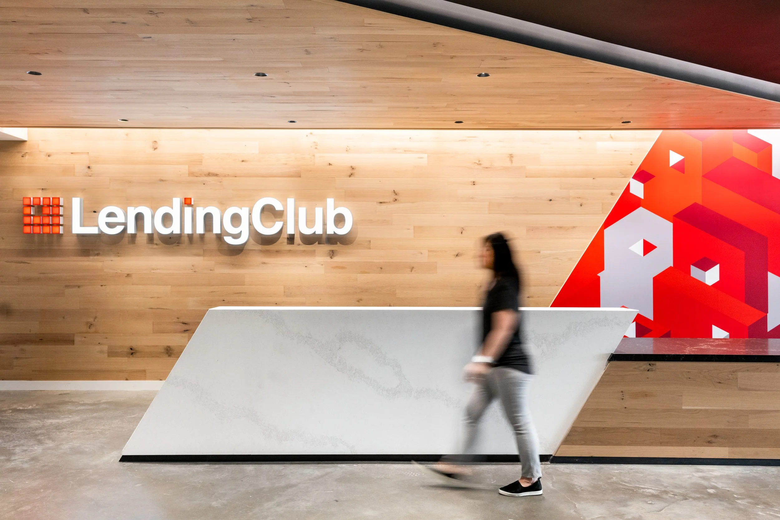 Interior of a modern office or lobby with a wooden wall featuring a illuminated 'LendingClub' sign and a colorful abstract geometric mural. A person in dark clothing is walking past a white reception desk.