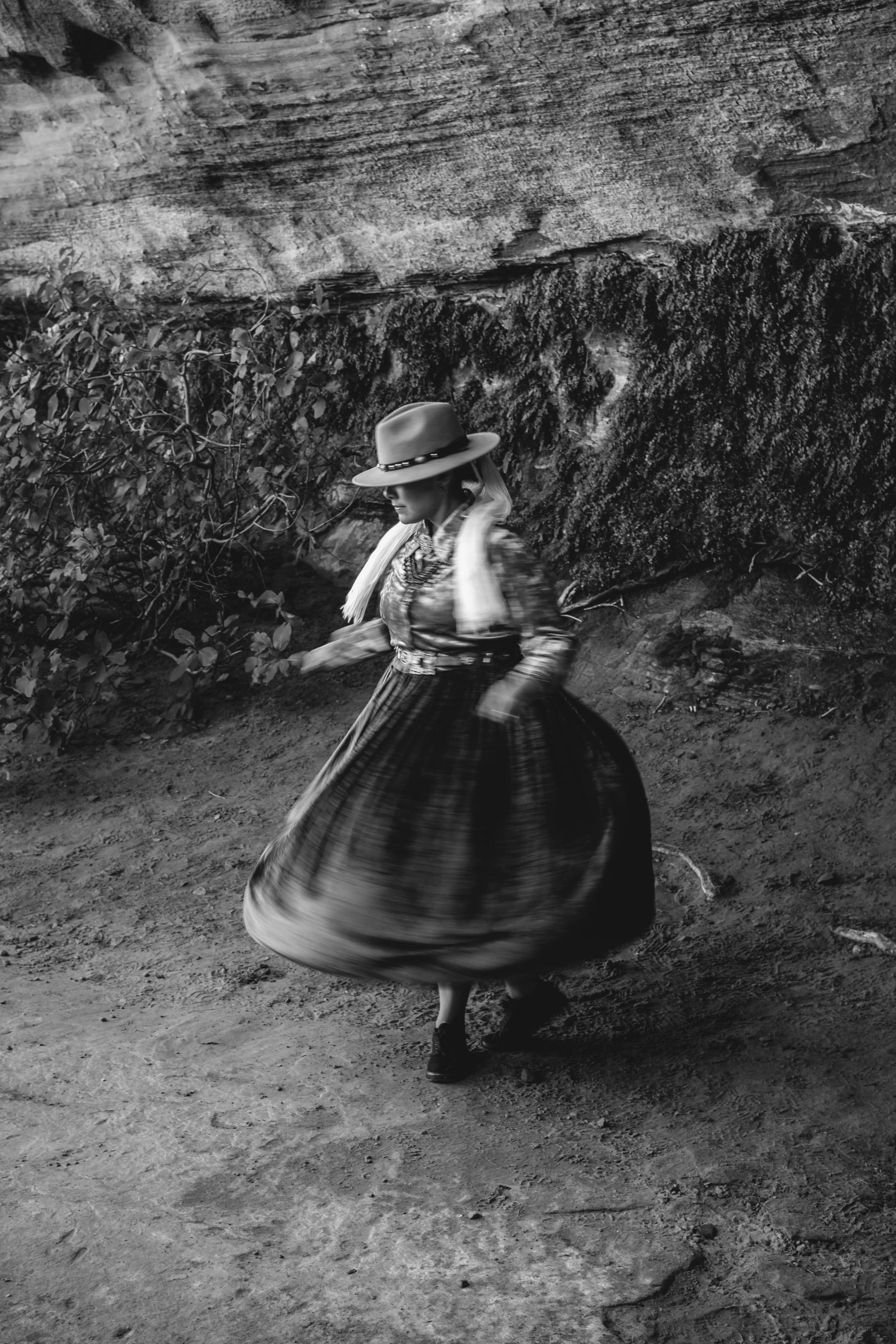 Black and white photo of a woman in a long dress and large hat twirling outdoors near a rocky cliff and bushes.