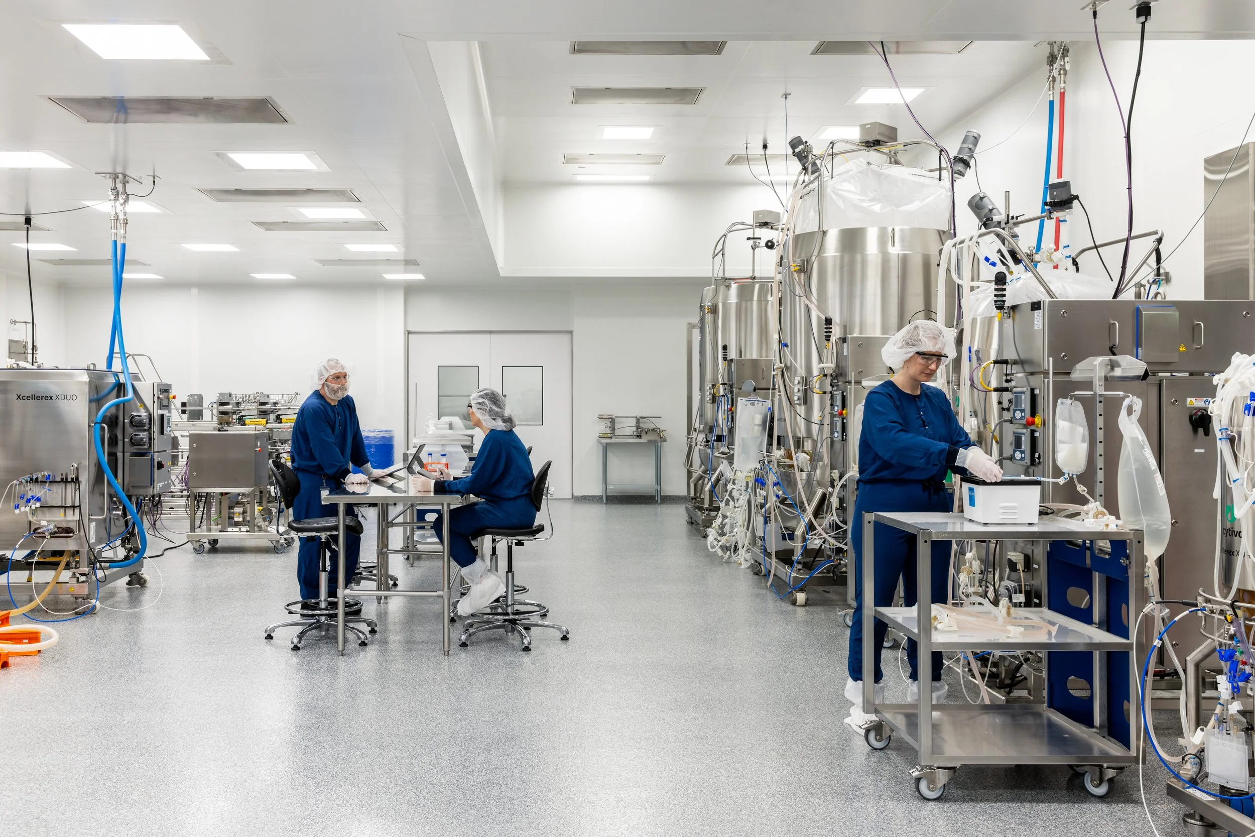 Three workers in a clean, sterile laboratory room working with scientific equipment, dressed in blue lab coats, hair coverings, and gloves.