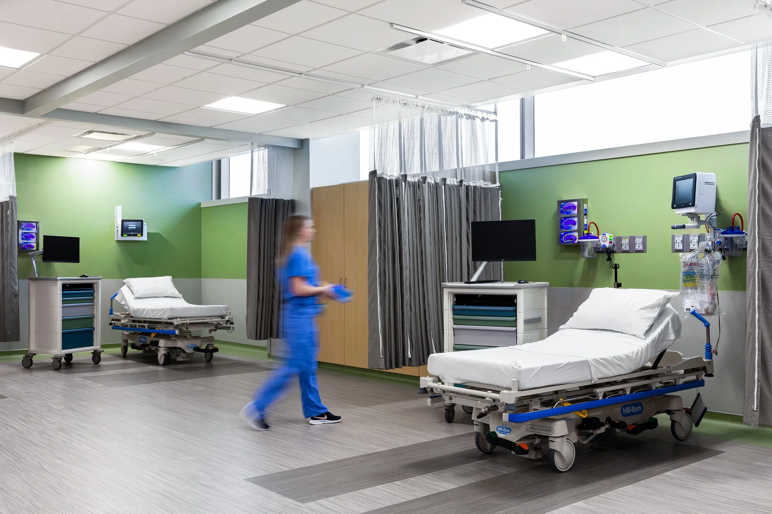 Hospital room with two empty beds, medical equipment, and a nurse walking past. The room has green walls, curtains separating beds, and medical monitors on the walls.