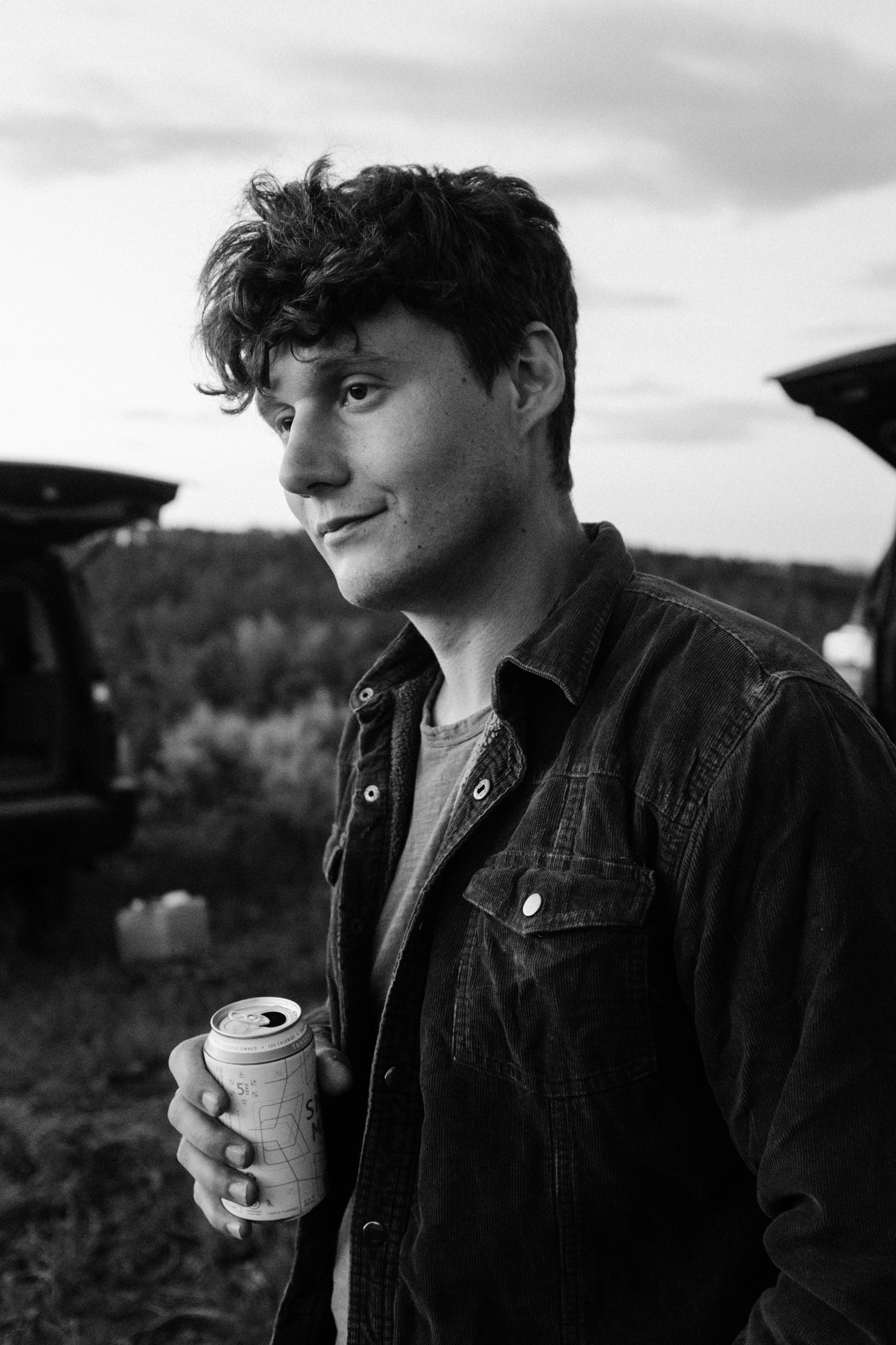 A young man with curly hair holding a can, standing outdoors with open car trunks behind him on a cloudy day.
