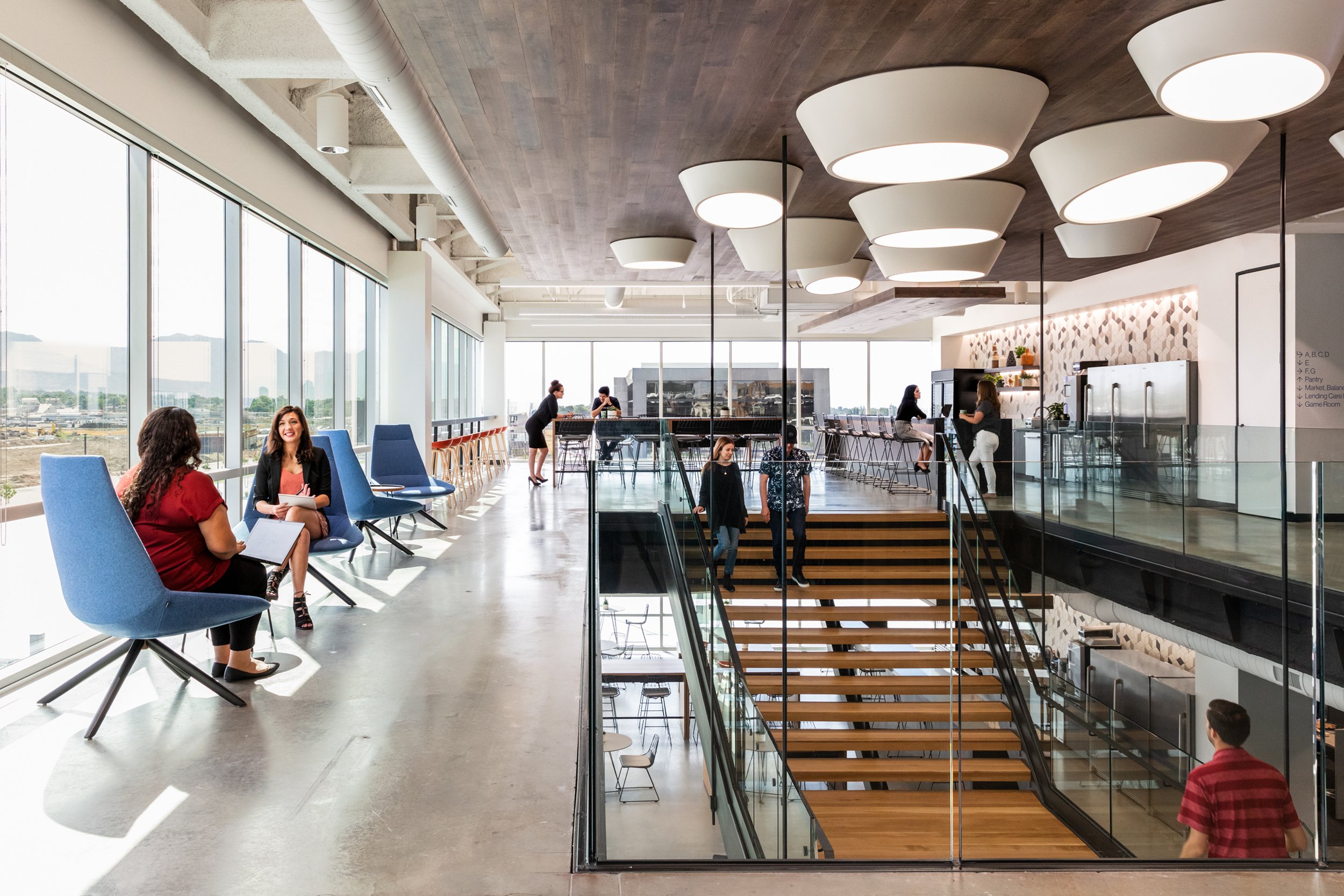 Interior of a modern office or coworking space with large windows, blue chairs, and people working and socializing near the windows and at a bar-height counter.