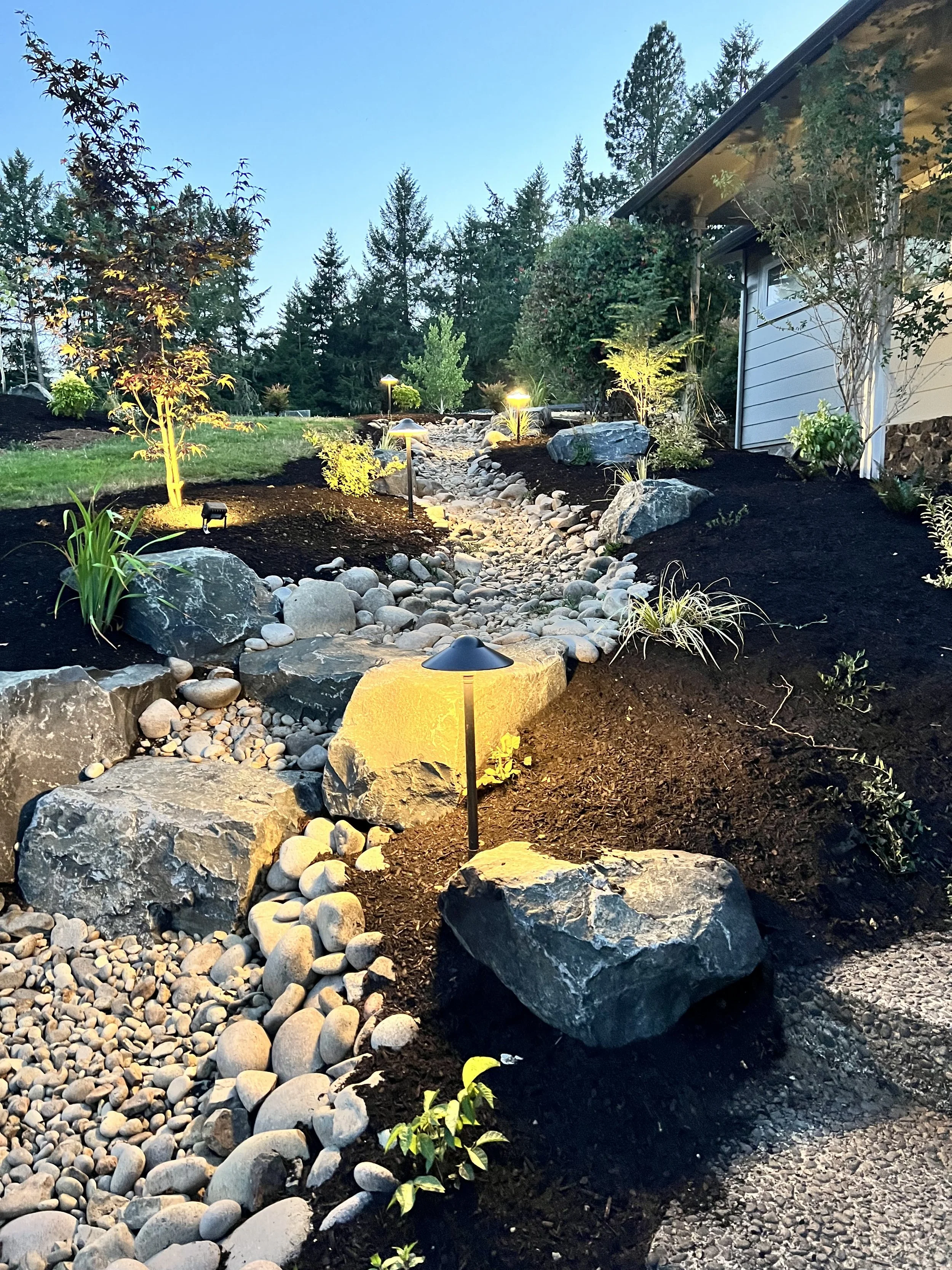 Well-lit landscaped garden with rocks, plants, and a winding stone pathway at dusk.