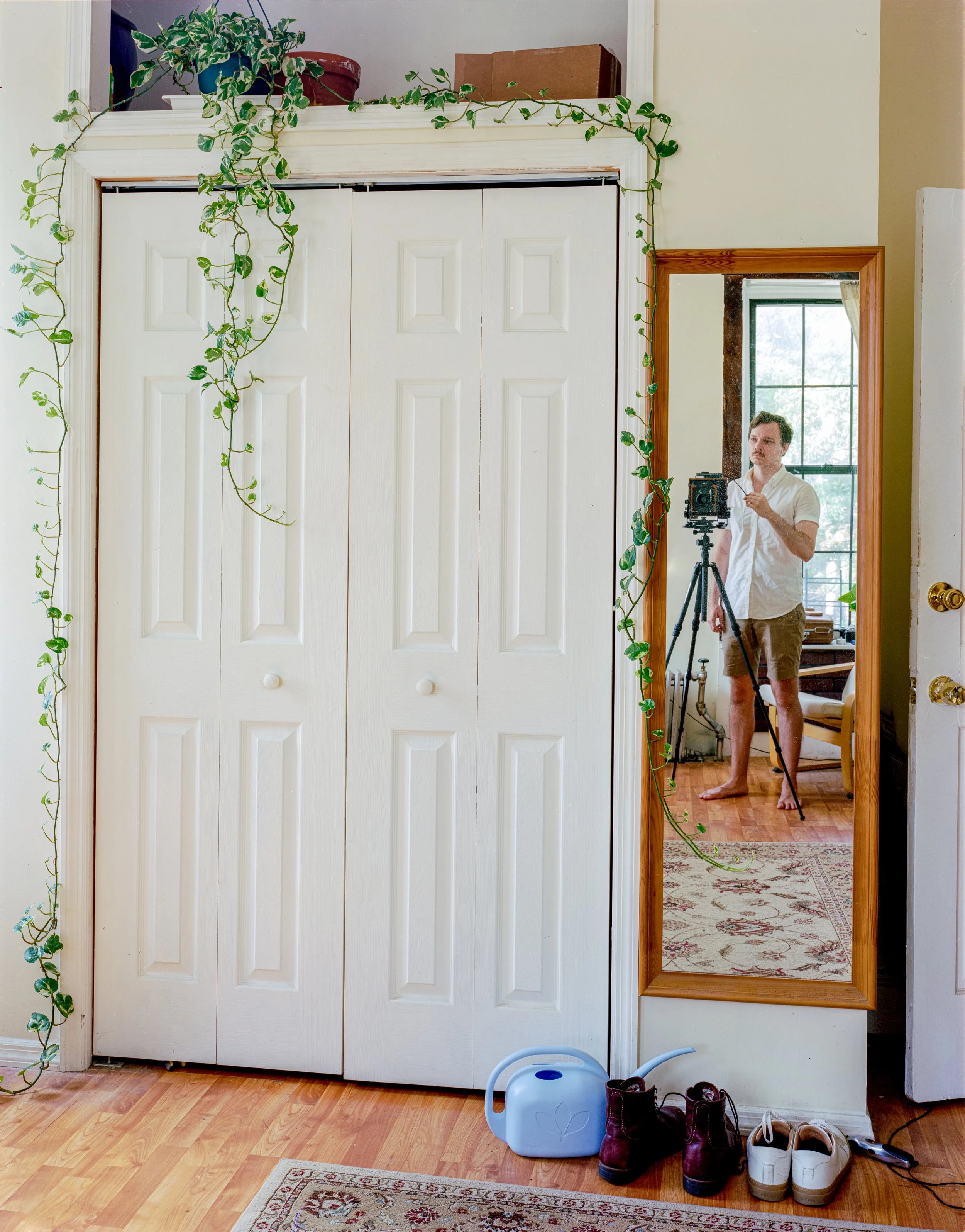 A person taking a mirror selfie with a camera on a tripod in a room. The room has a closet with closed white bi-fold doors, a large window, a plant with green vines hanging around the mirror frame, and some shoes and a watering can on the floor.