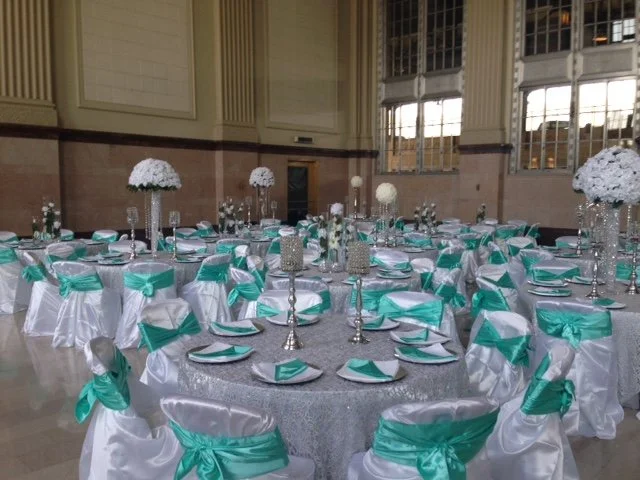 Wedding reception setup at T&P Station ballroom featuring silver sequin tablecloths, white chair covers with teal satin bows, tall floral centerpieces, and crystal candle holders beneath the grand Art Deco windows.