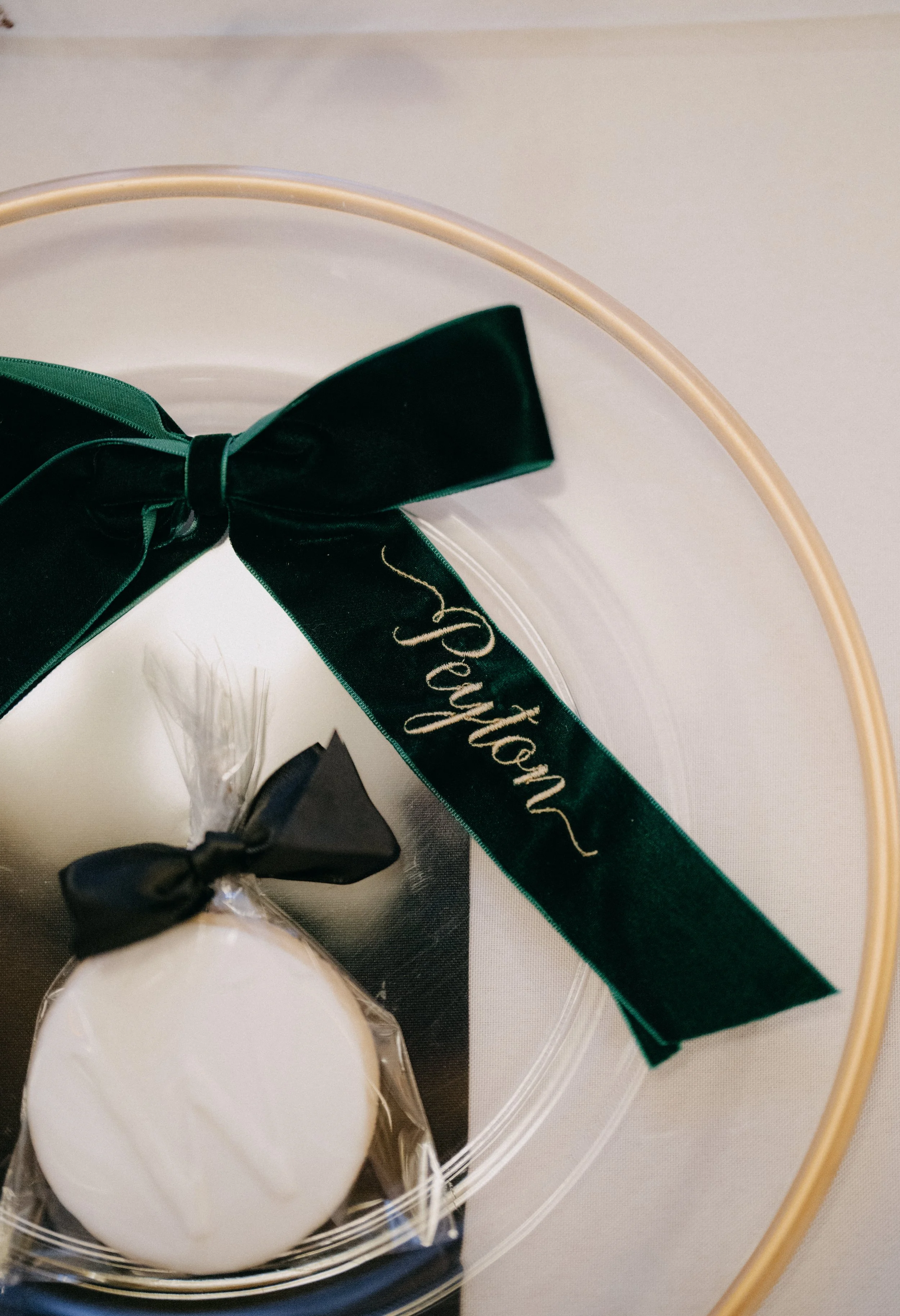 Elegant place setting with a green velvet ribbon name tag reading “Peyton” and a white favor cookie on a clear glass charger accented with gold trim.