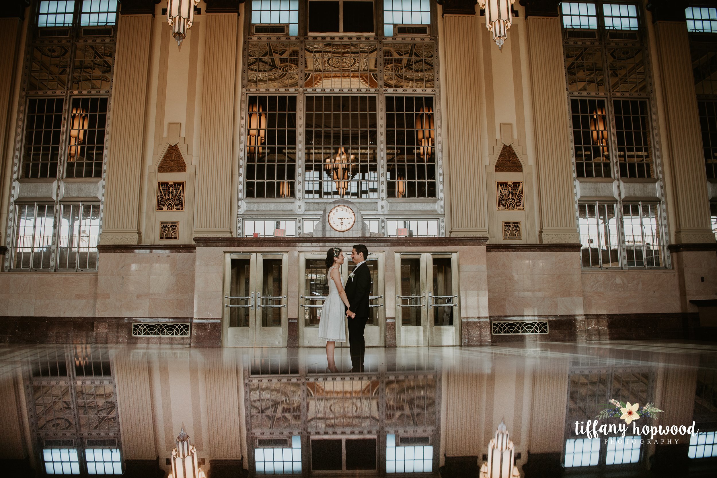 A bride and groom stand facing each other in a large, ornate train station with high ceilings, tall windows, and art deco design. Their reflection is seen in the shiny floor below them.