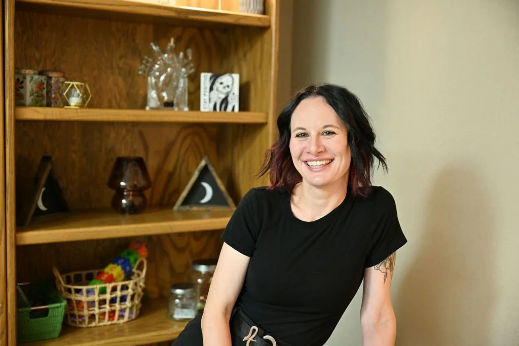 A woman with black and purple hair, wearing a black t-shirt, smiling and leaning against a wall in front of a wooden bookshelf with decorative items and children's toys.