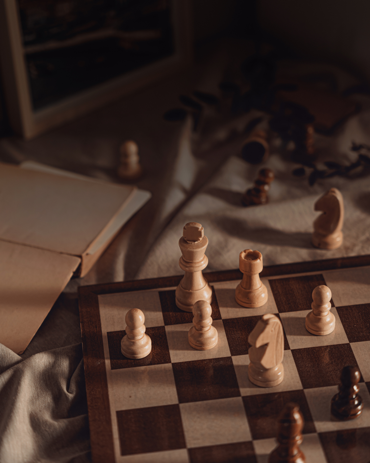 A chessboard with wooden chess pieces, some captured and scattered nearby, with an open book and a chess set in the background.