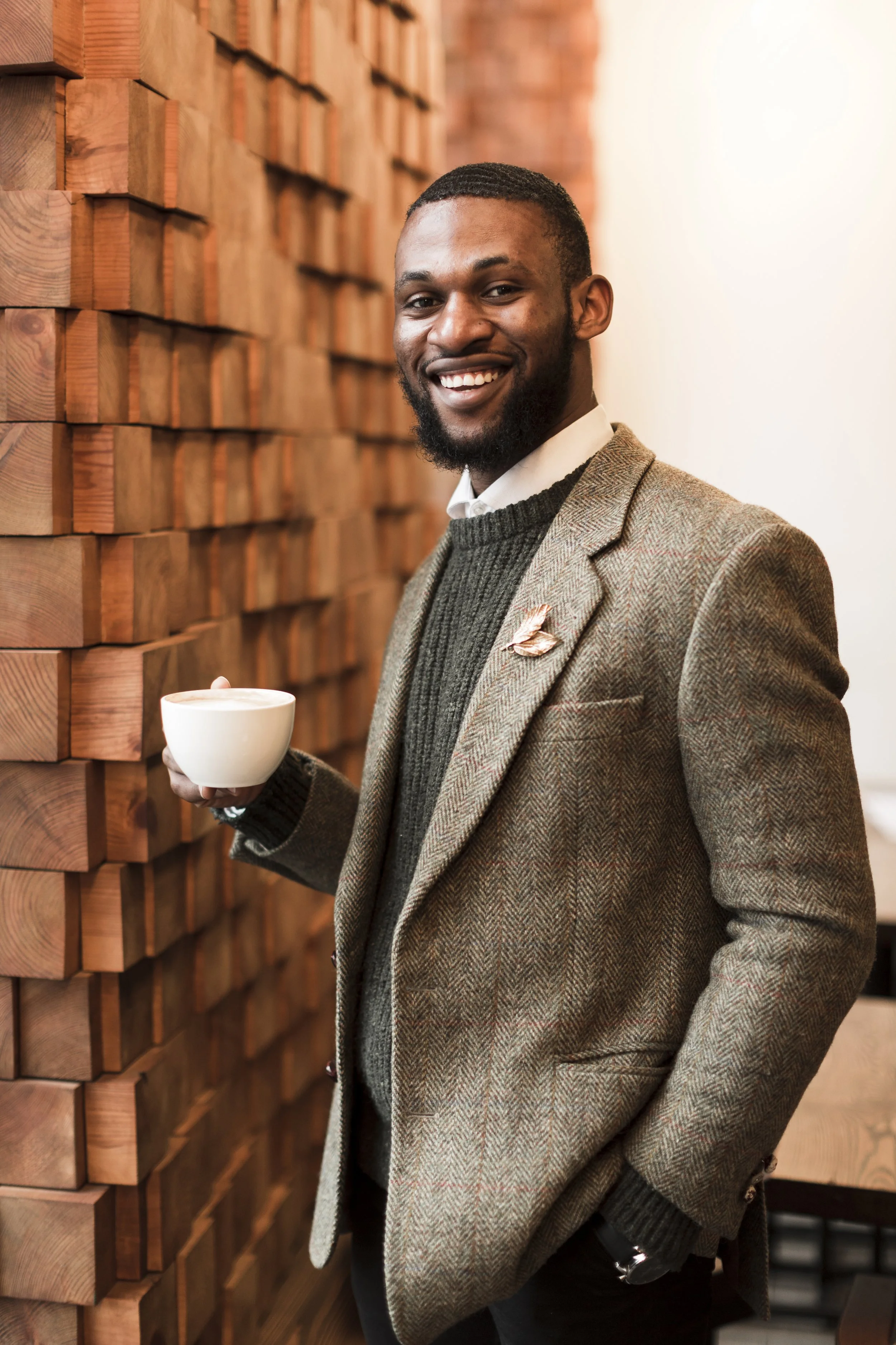 Un homme souriant portant un costume en tweed, tenant une tasse dans un café avec un mur en bois derrière.