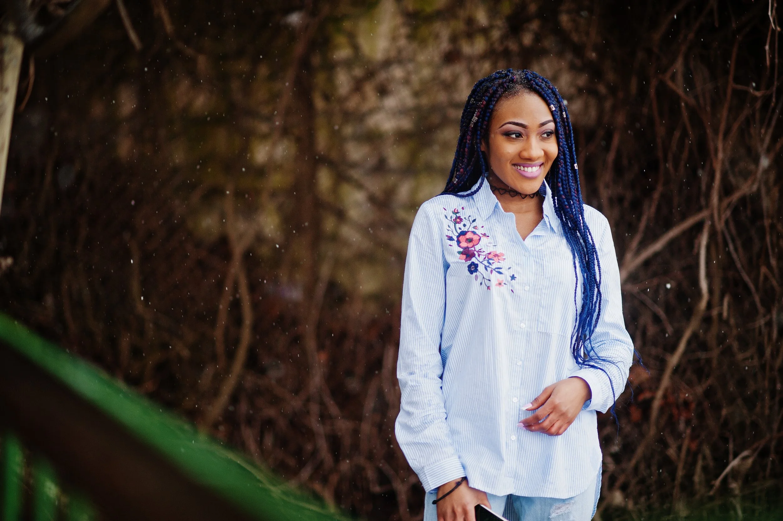 Une jeune femme souriante avec des cheveux en tresses bleues, portant une chemise blanche à broderie florale, debout devant un arbre.
