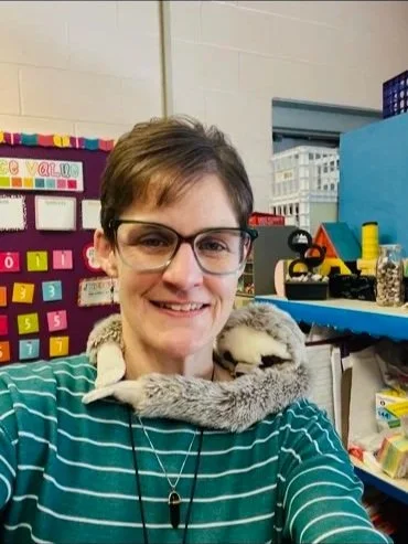 A woman with short brown hair and glasses smiling at the camera, wearing a teal and white striped shirt, with a gray and white ferret on her shoulder in a colorful classroom setting.