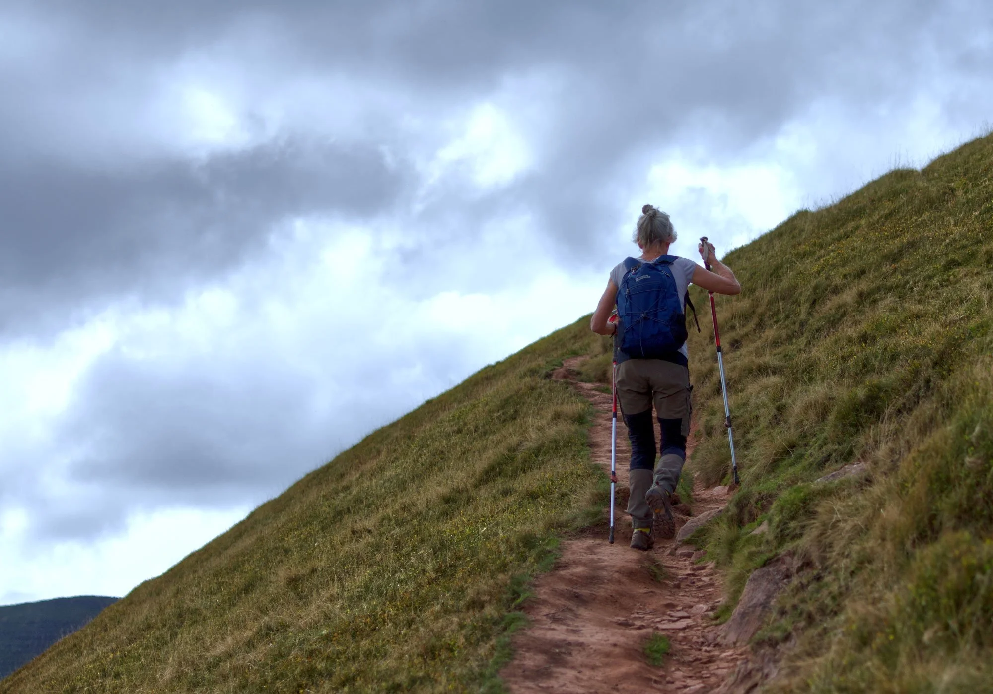 Hiker walking uphill on a dirt trail surrounded by grassy hill, carrying a blue backpack and using trekking poles, under cloudy sky.