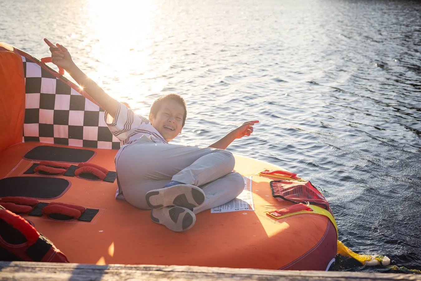A young boy lying on an orange inflatable boat on the water, smiling and pointing, with the sun setting in the background.