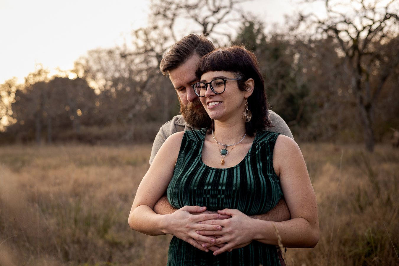 A couple embraces in a field during sunset, with the man supporting the woman from behind.