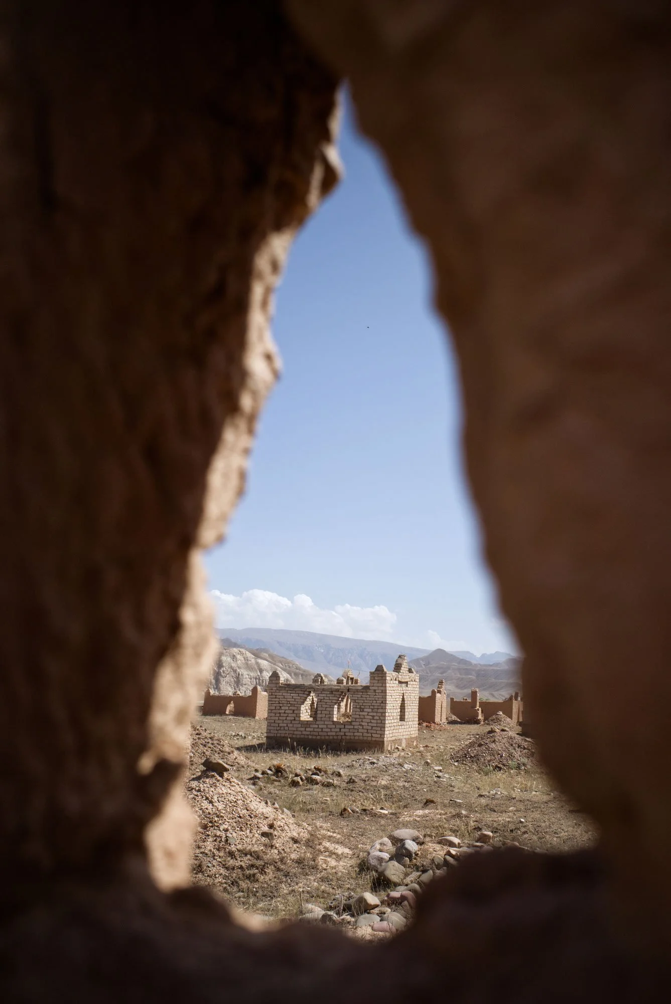 Ruins of an adobe building seen through a small hole in a wall in a desert landscape with mountains in the background.