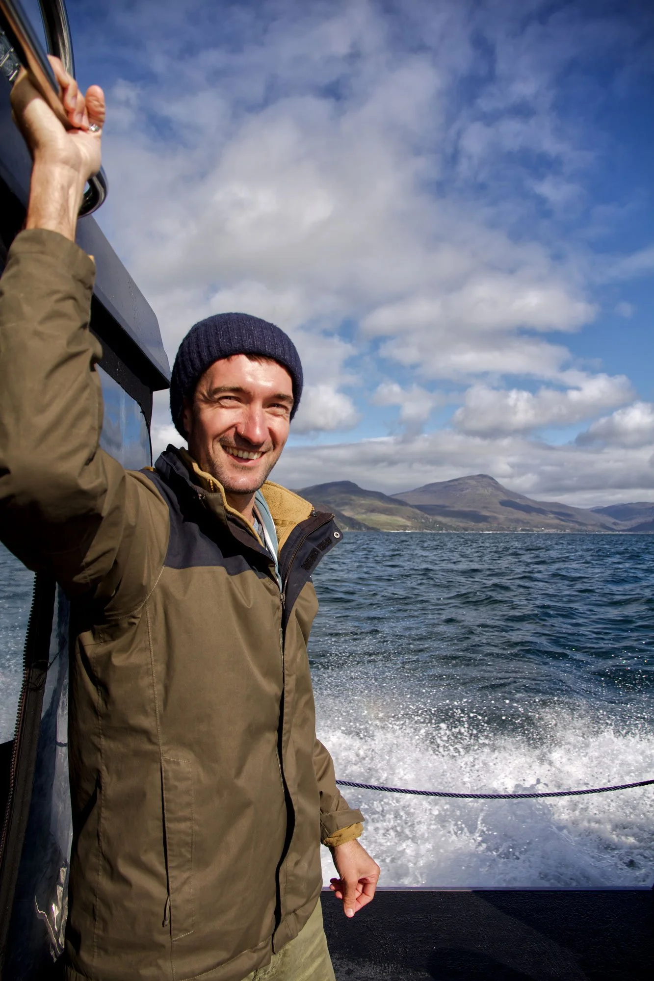 Smiling man in a brown jacket and navy beanie on boat with water and mountains in the background
