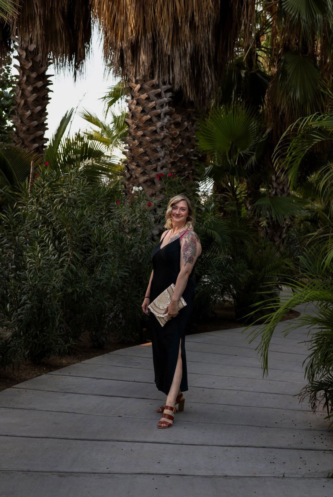A woman in a black dress with a high slit, tan heeled sandals, and holding a textured clutch, stands on a curved pathway surrounded by lush green tropical plants and tall palm trees.