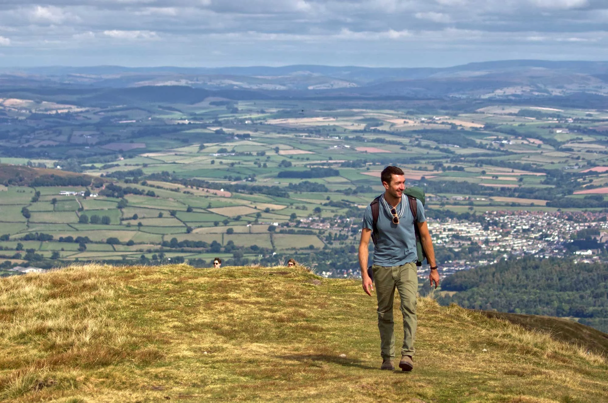 A man in outdoor hiking clothes walking on a grassy hilltop overlooking a large, patchwork landscape of farmland and small towns in the distance.