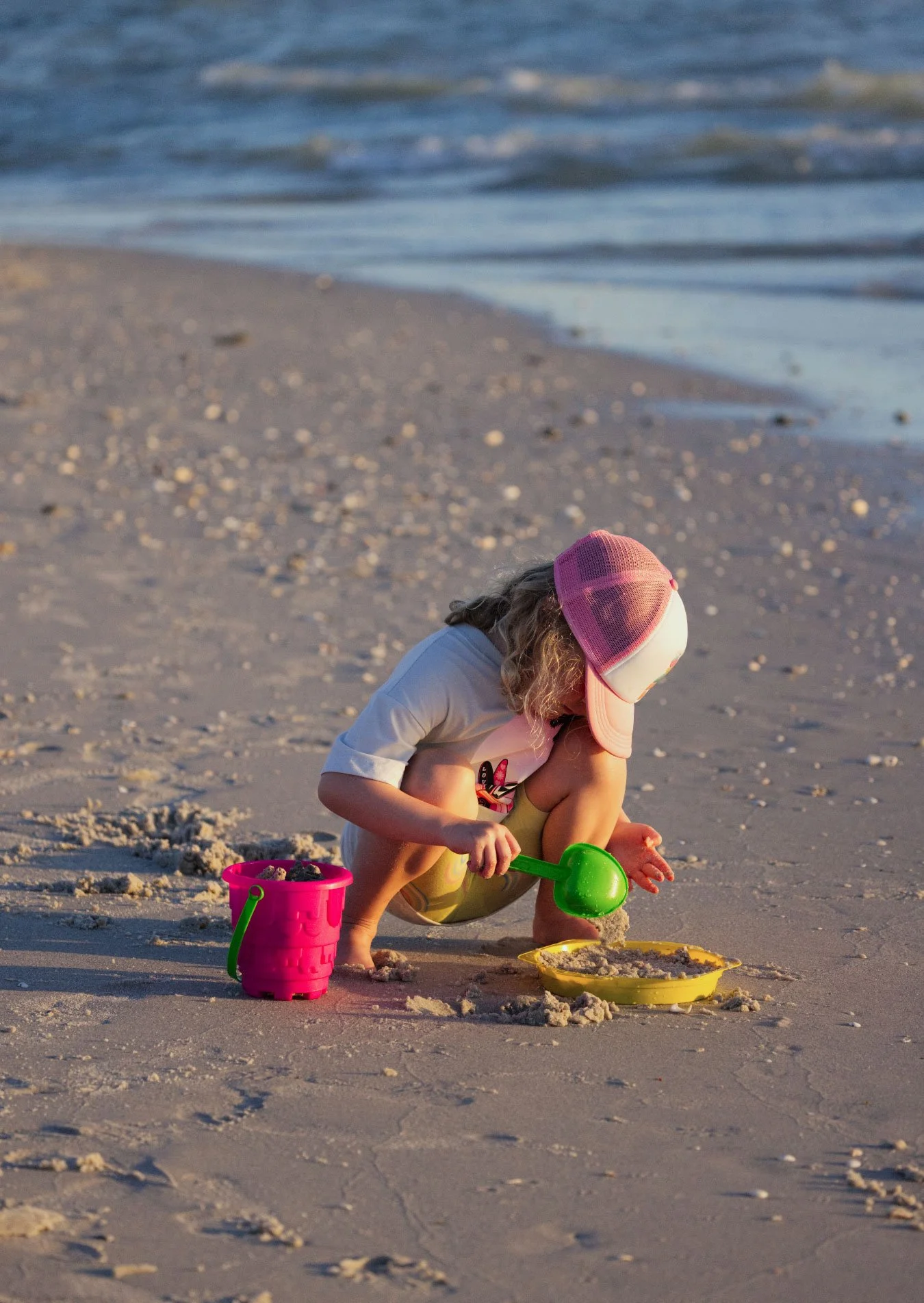 Child wearing a pink cap and white shirt playing with sand on the beach using a green shovel, with a pink bucket and yellow tray nearby.