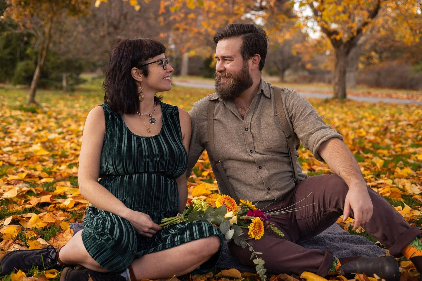 A couple sitting on a blanket in a park filled with fallen autumn leaves, looking at each other and smiling, with trees displaying fall foliage in the background. The woman is holding a bouquet of flowers.